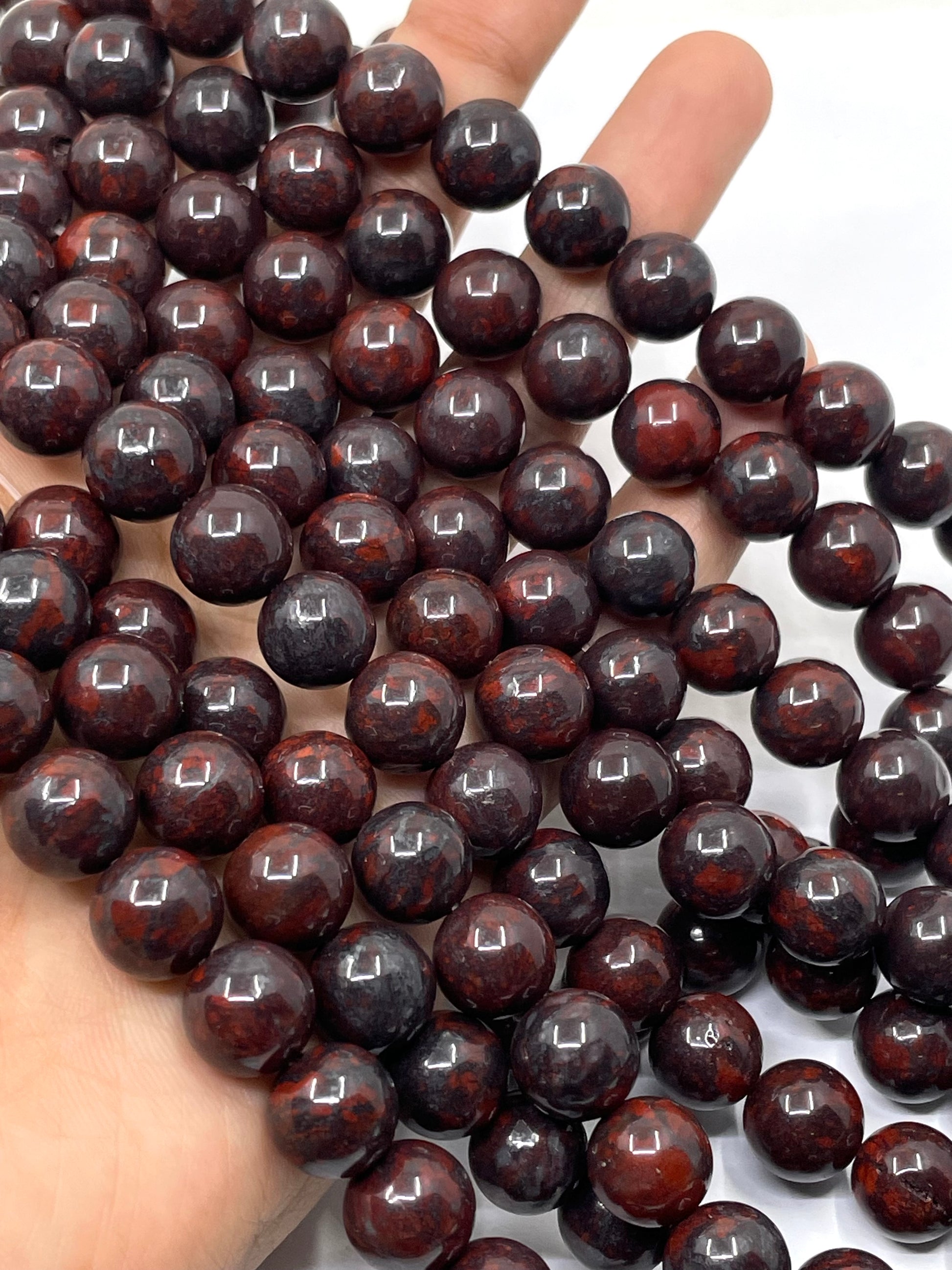 A close-up of glossy dark reddish-brown round brecciated jasper beads draped over a hand, with the beads filling most of the frame against a white background.