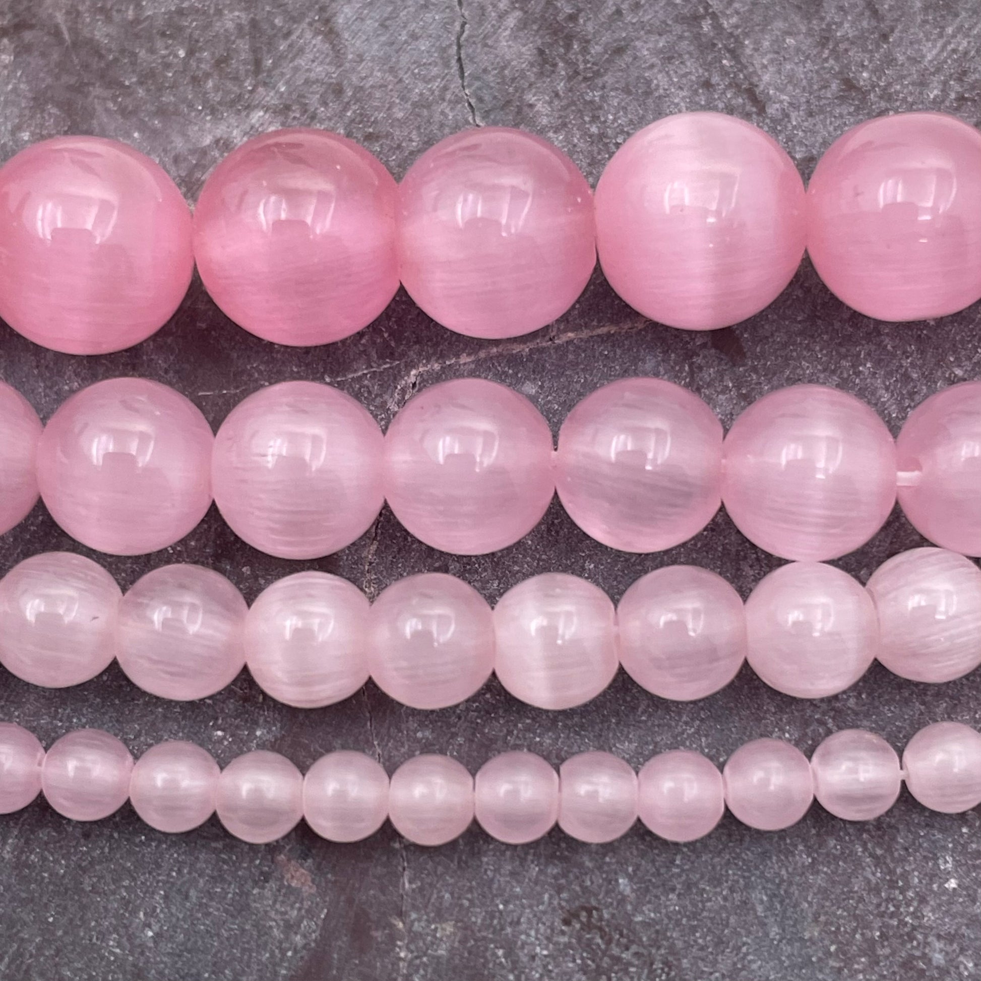 Four strands of pink cats eye round gemstone beads in varying sizes, arranged horizontally on a textured gray surface.