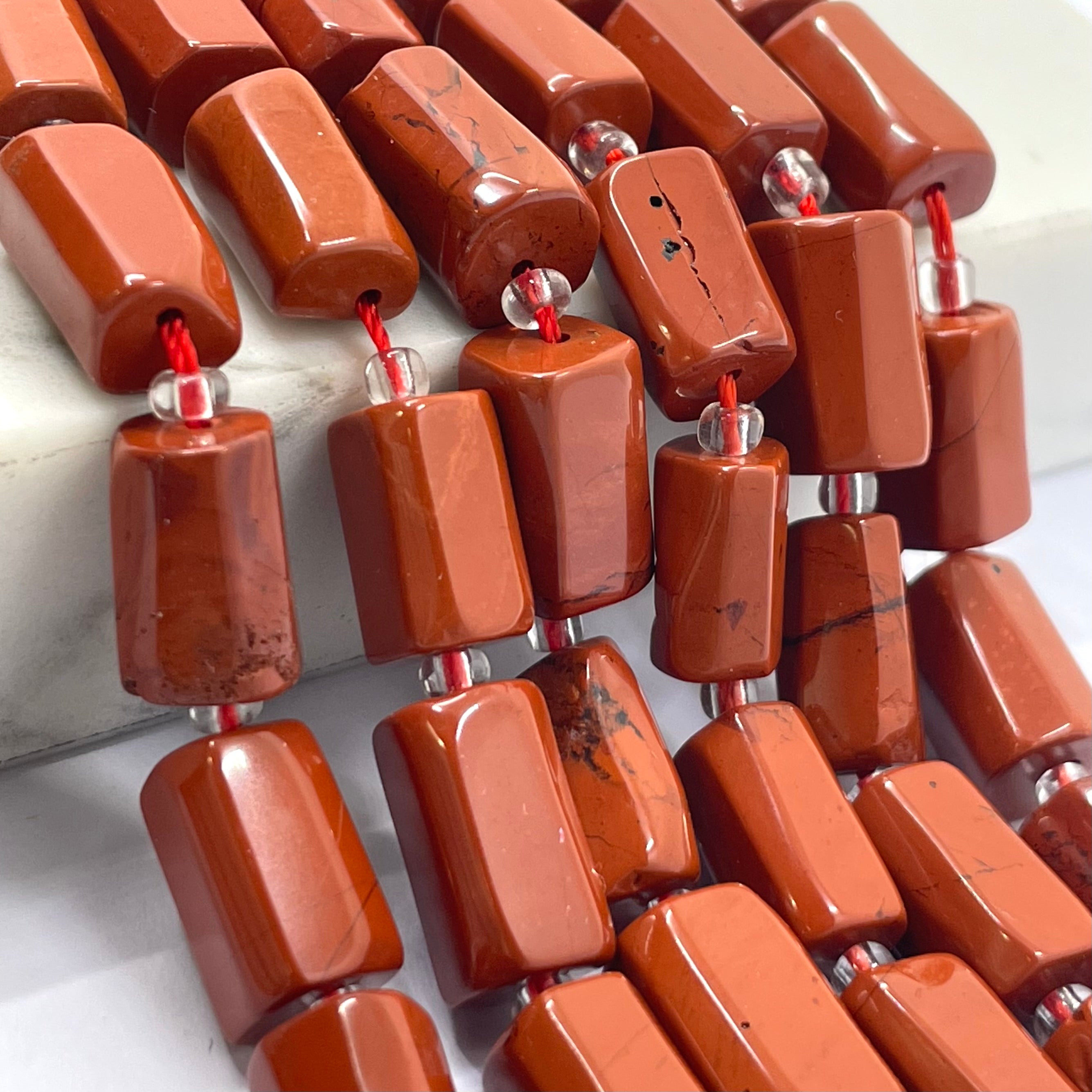 Rectangular red jasper beads with a polished, glossy finish, threaded on red cords with small silver spacer beads, shown in multiple strands on a light background.