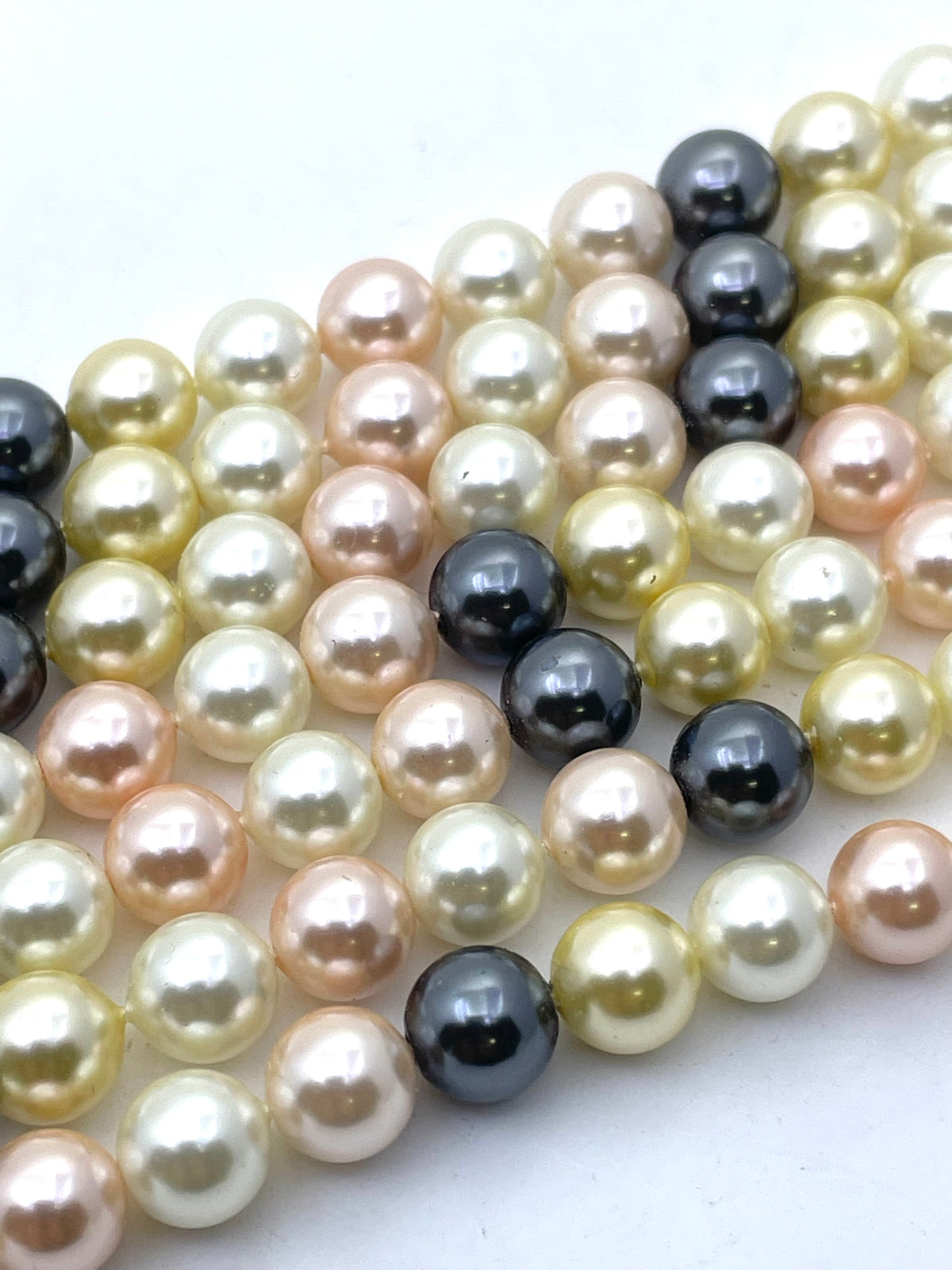 A close-up of strands of shell pearl beads in mixed colors including cream, peach, champagne, and dark charcoal black, arranged diagonally on a plain white background.