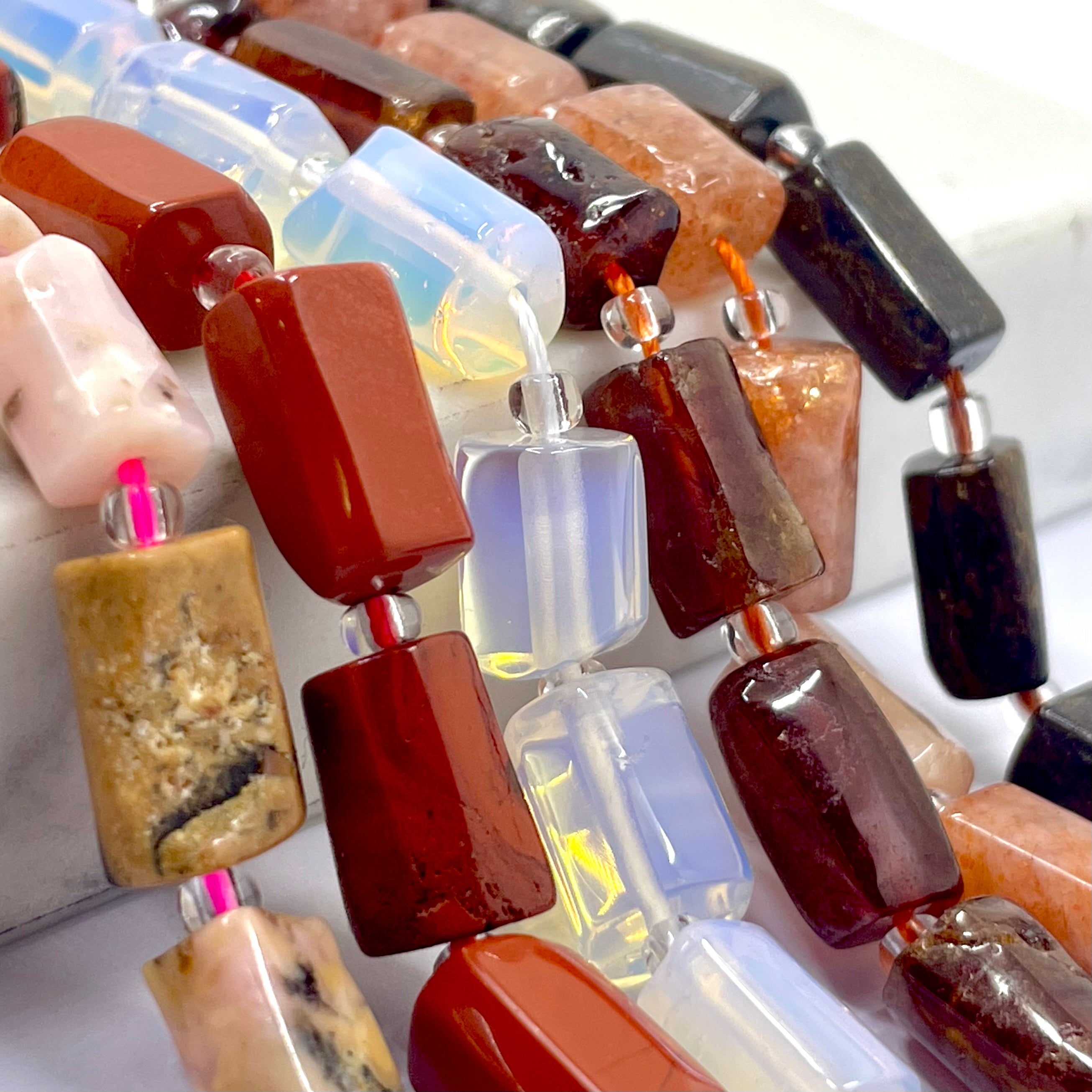 Close-up of a beaded strand featuring polished rectangular gemstone beads in rich reds, deep browns, soft pinks, translucent milky whites, and a few dark near-black pieces, separated by small silver spacers.