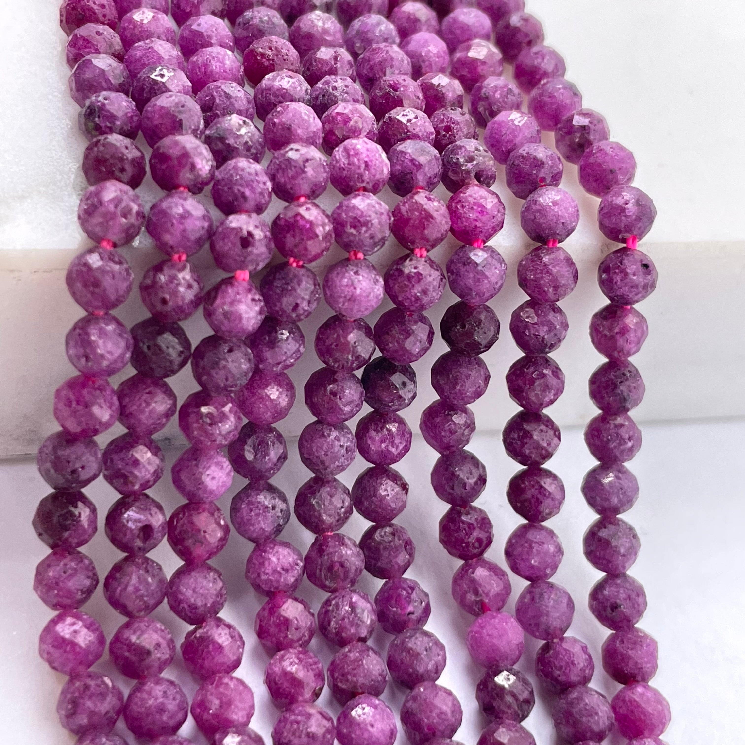 Multiple strands of faceted ruby gemstone beads, draped vertically on a white surface.