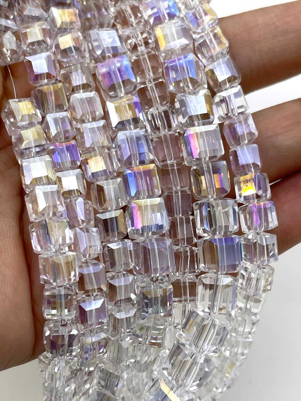 Clear cube faceted beads with an iridescent AB finish, draped over a hand to show their transparent and rainbow-reflective quality.