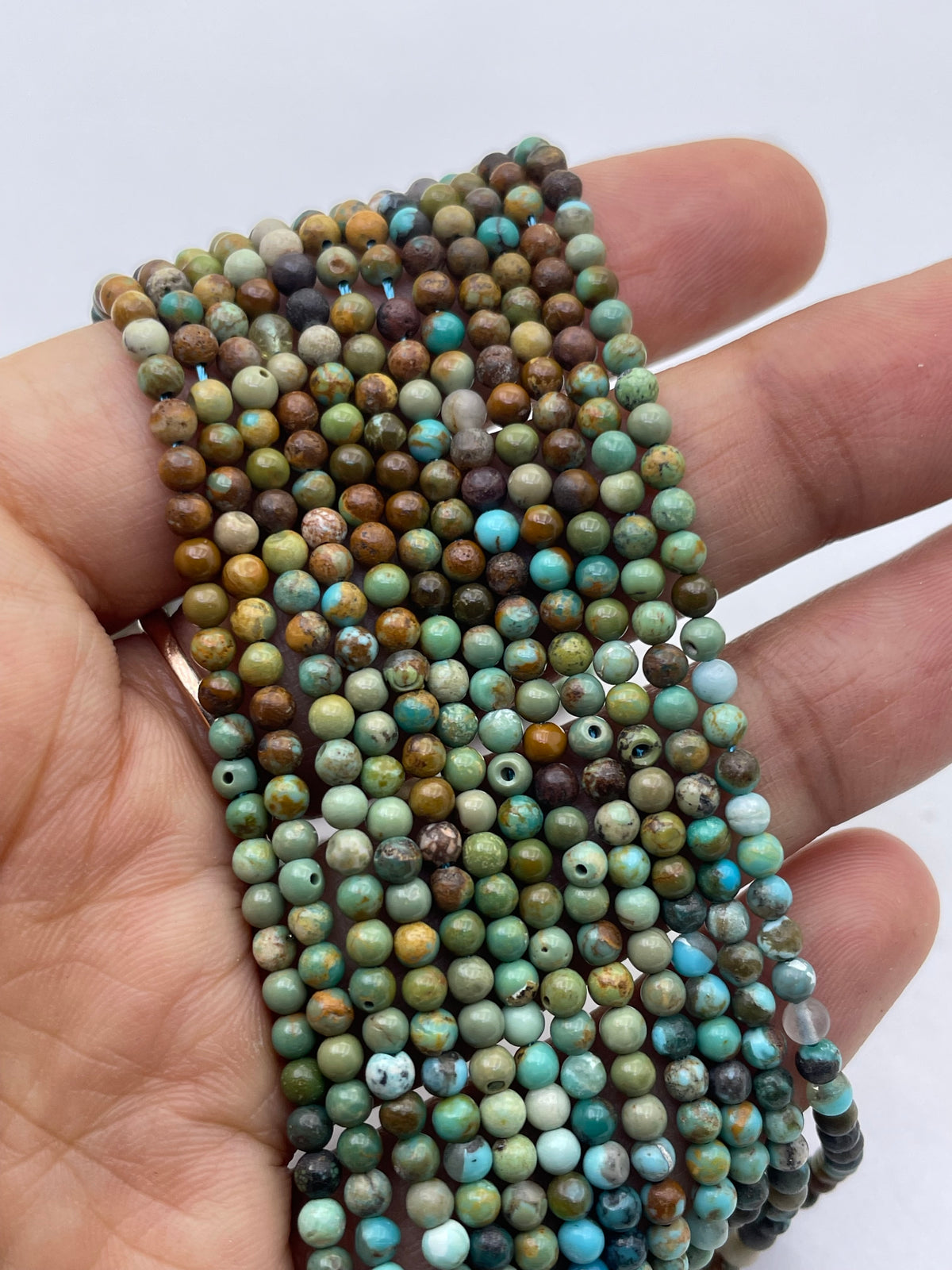 A hand holding a bundle of small round turquoise beads in mixed earthy and turquoise colors, including green, blue, brown, beige, and white, against a plain white background.