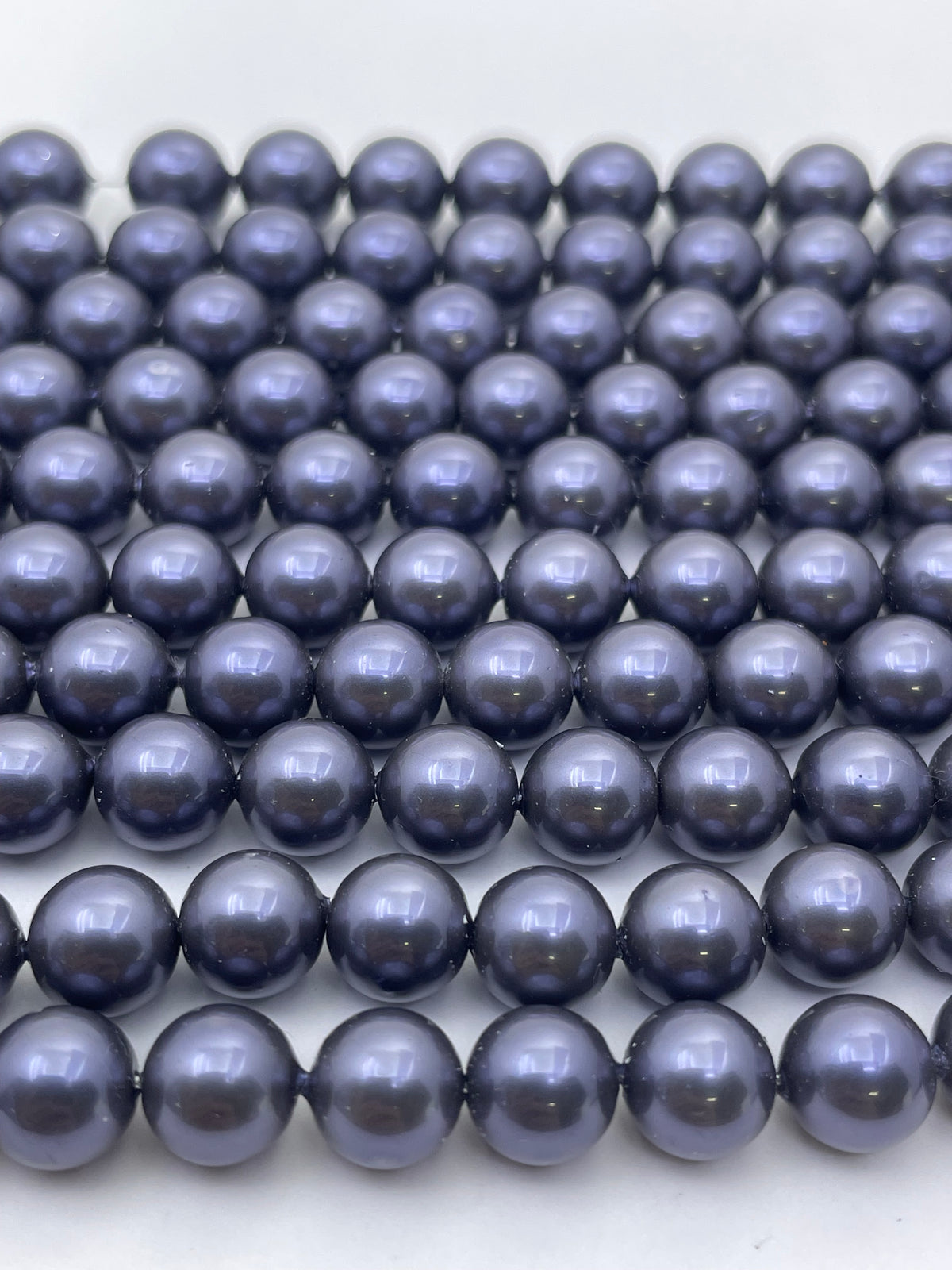A close-up of multiple strands of glossy, dark grayish-blue shell pearl beads arranged in neat rows on a plain white background.