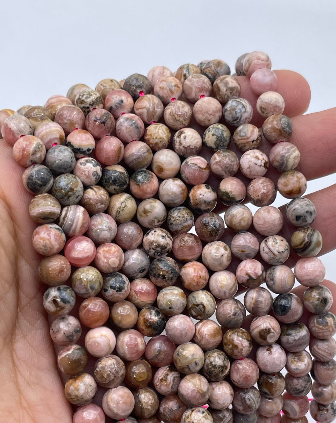 A hand holding a bundle of round, polished pink and brown rhodochrosite stone beads with natural marbled patterns, set against a plain light gray background.