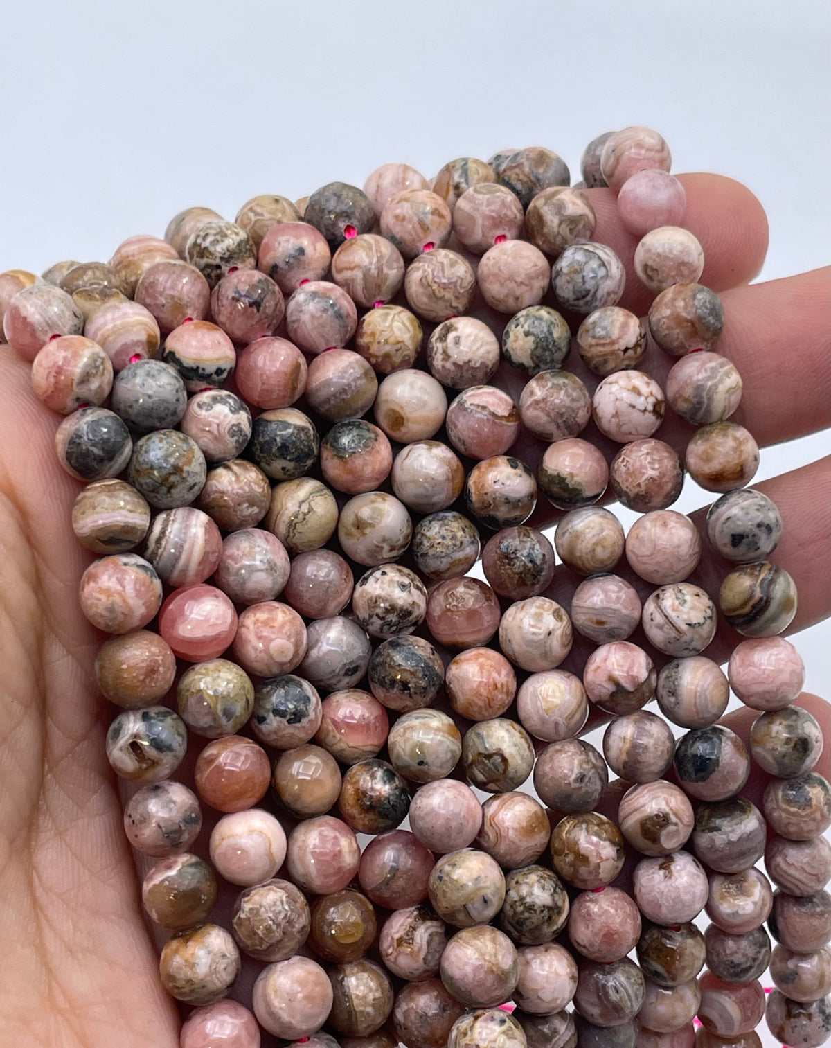 A hand holding a bundle of round, polished pink and brown rhodochrosite stone beads with natural marbled patterns, set against a plain light gray background.