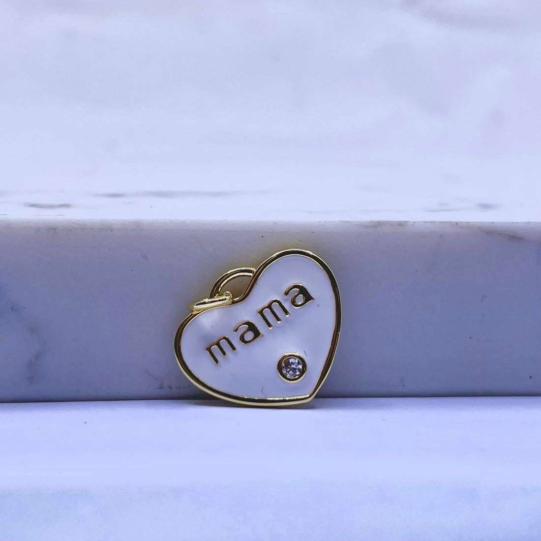 A small heart-shaped pendant charm with a gold border, white enamel fill, the word 'mama' in gold lettering, and a small clear rhinestone, resting on a white marble block.