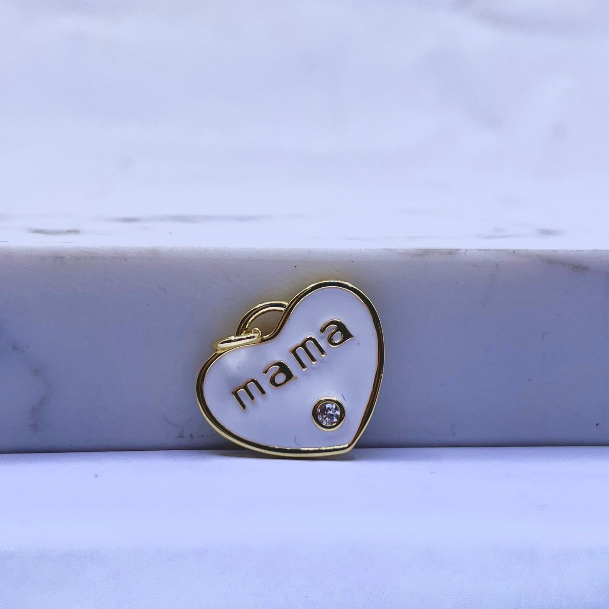 A small heart-shaped pendant charm with a gold border, white enamel fill, the word 'mama' in gold lettering, and a small clear rhinestone, resting on a white marble block.