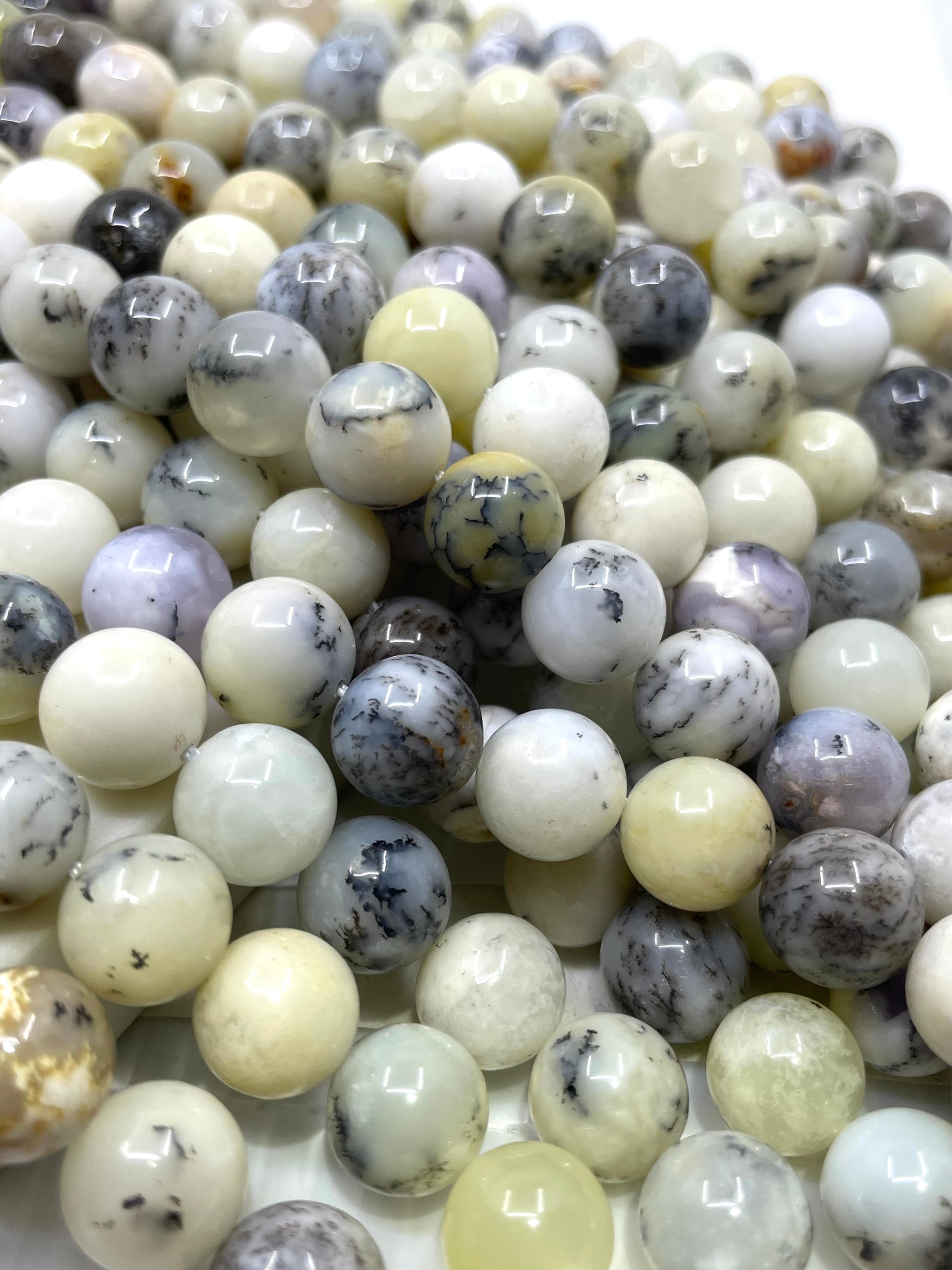 A close-up pile of polished round gemstone beads in mixed colors including cream, white, gray, lavender, and yellowish tones, with some beads showing natural marbling or inclusions.