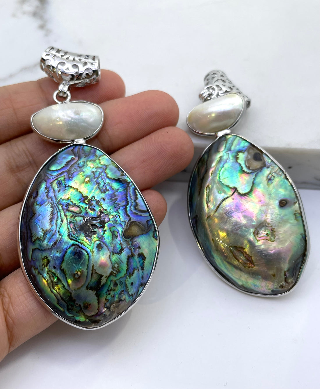 Teardrop-shaped abalone shell pendant inlays with iridescent blue, green, and purple hues, accented by small white pearls and textured silver posts, displayed on a white marble surface.