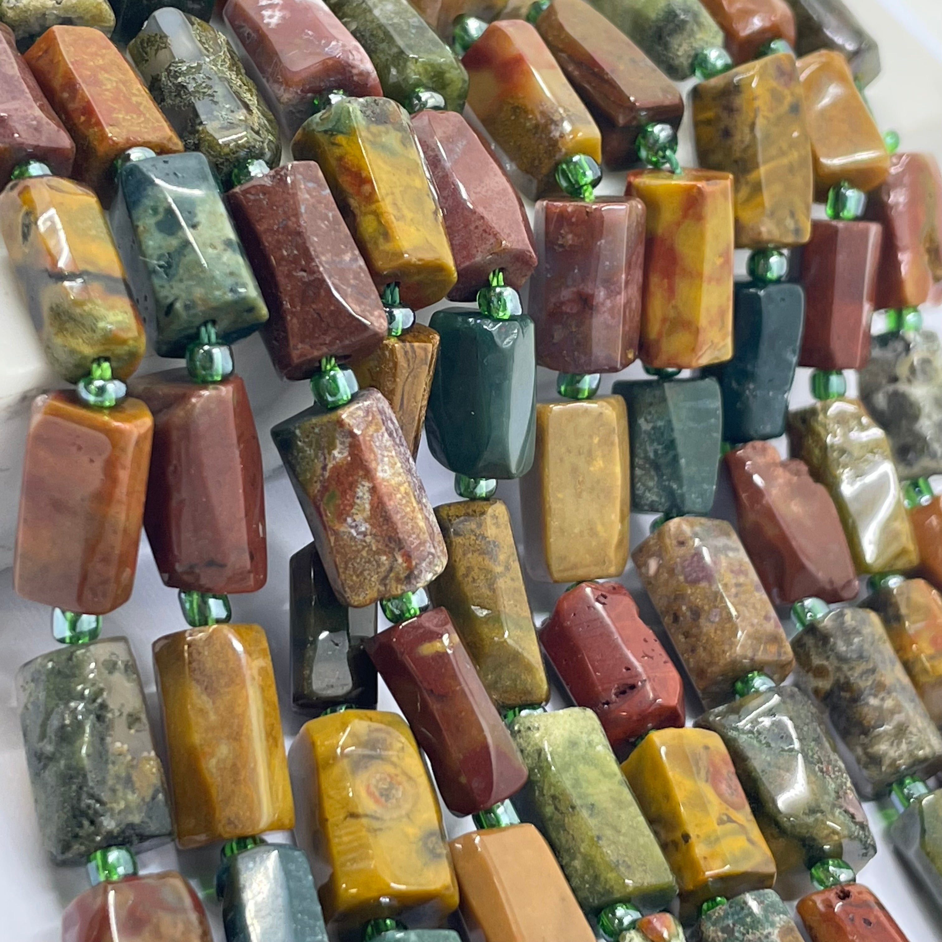 A close-up of multiple strands of polished rectangular ocean agate gemstone beads threaded on green cord. The beads display a wide variety of natural stone patterns and colors, including yellow, red, green, brown, and gray, arranged in diagonal rows.