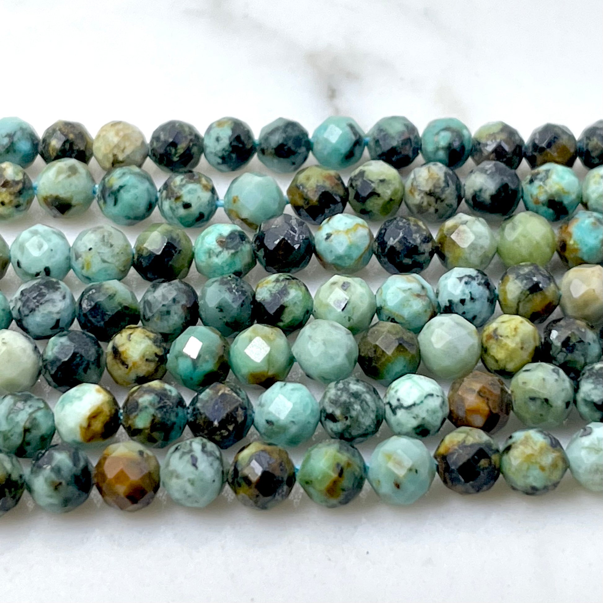Faceted round african turquoise gemstone beads in a marbled mix of turquoise, green, black, brown, and cream, arranged in rows on a white marble surface.