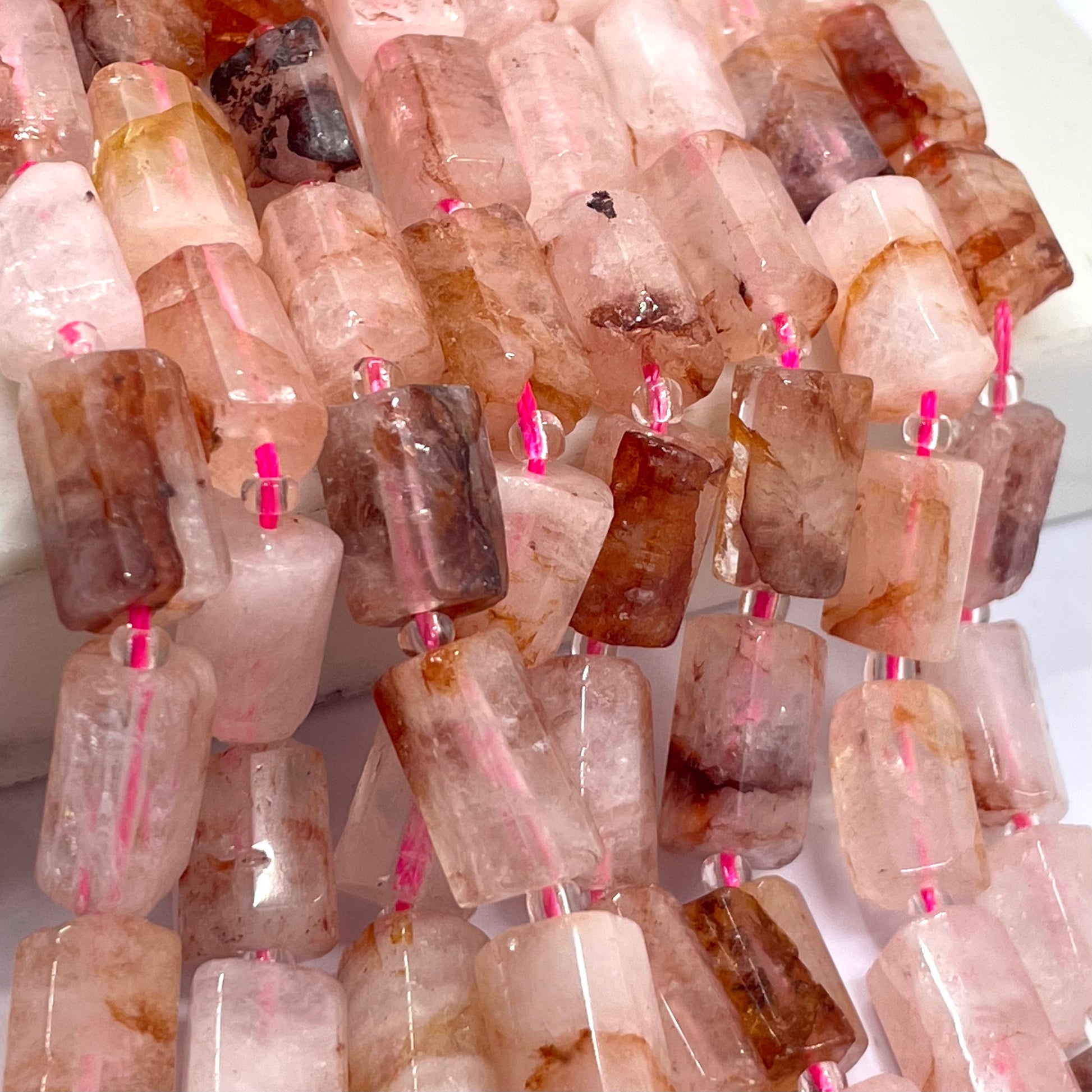 A close-up of a strand of polished rectangular fire quartz gemstone beads, showing natural pink, peach, and brown crystalline patterns, strung on a white cord with small pink loops.