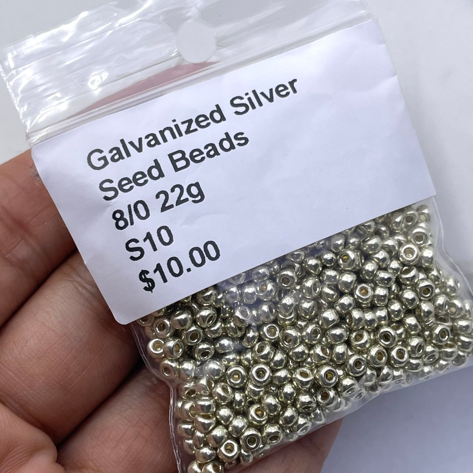 A hand holding a clear plastic bag filled with small metallic seed beads. A white label on the bag reads 'Galvanized Silver Seed Beads 8/0 22g S10 $10.