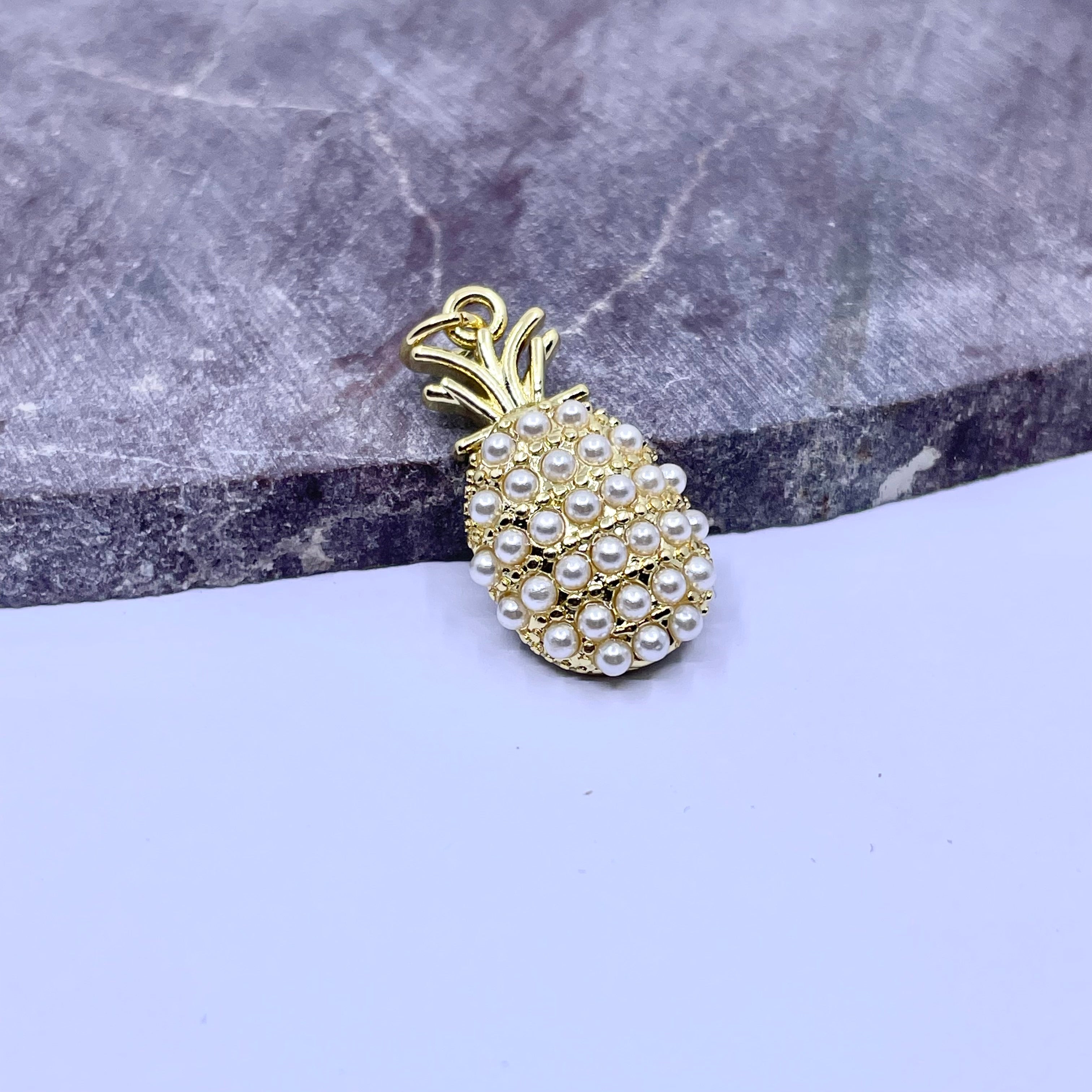 A pineapple-shaped jewelry charm or pendant with a gold-tone metal body and white faux pearls, resting on a split gray stone and light lavender surface.