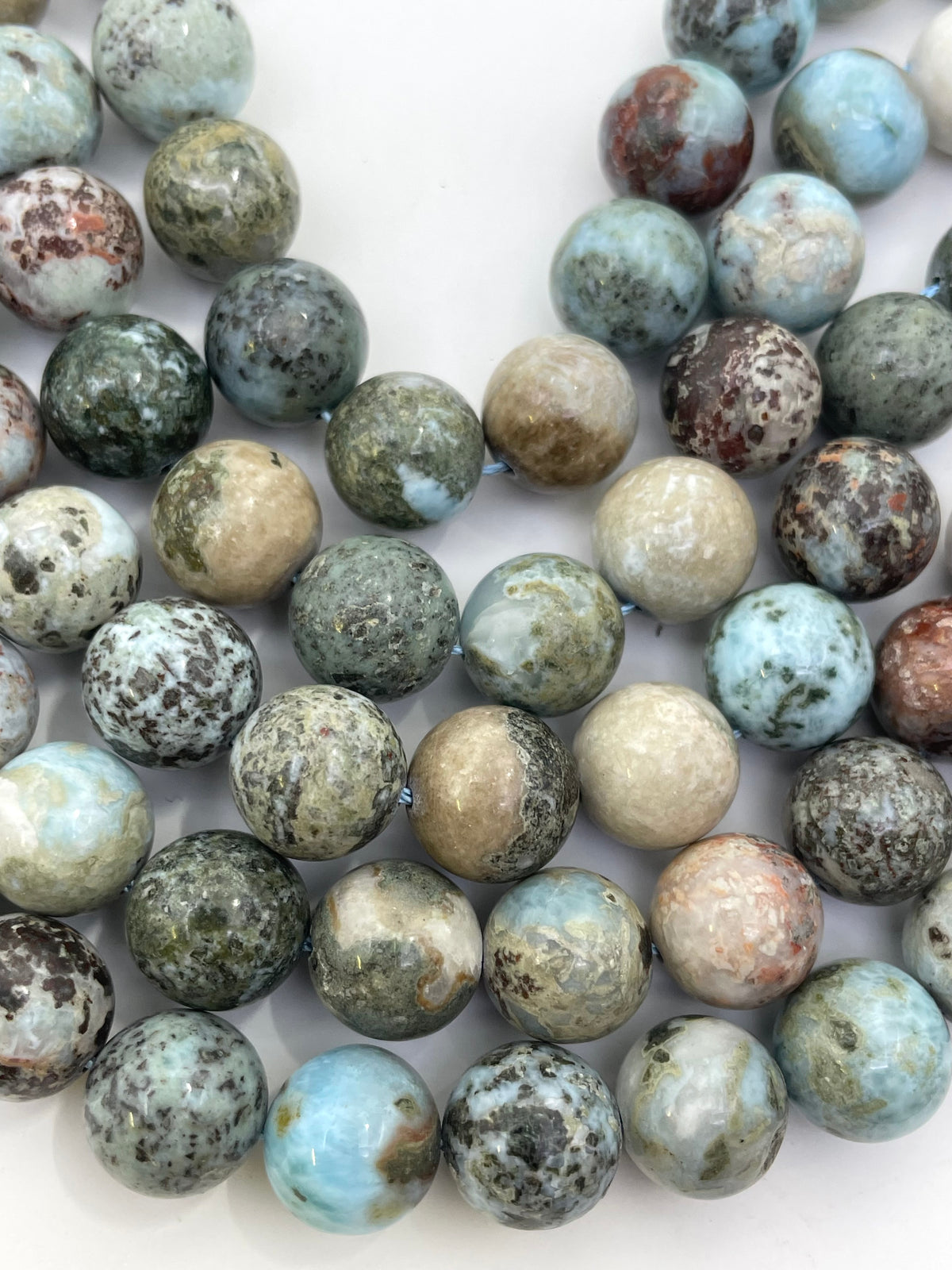 A close-up of round polished larimar stone beads in a mottled mix of light blue, gray, beige, green, and brown, arranged on a plain white background.