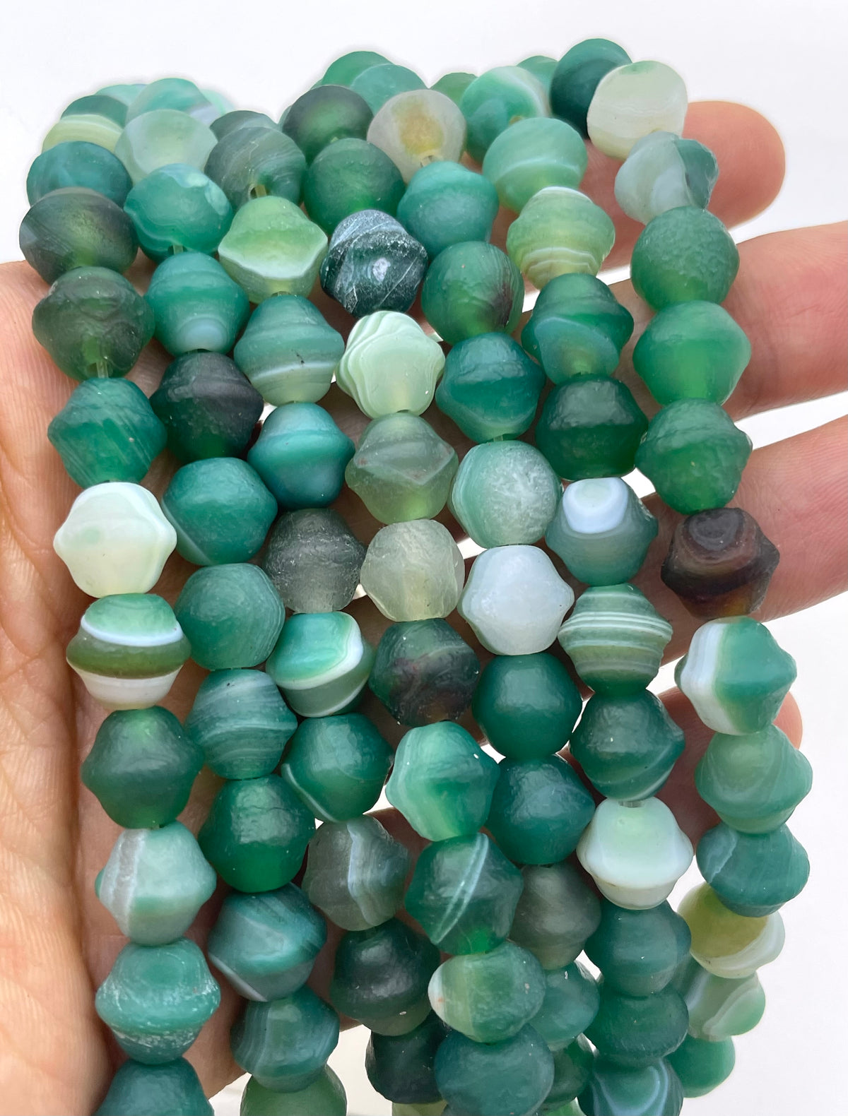 A hand holding multiple strands of faceted green agate stone beads, including shades of emerald, teal, and white marbled beads.