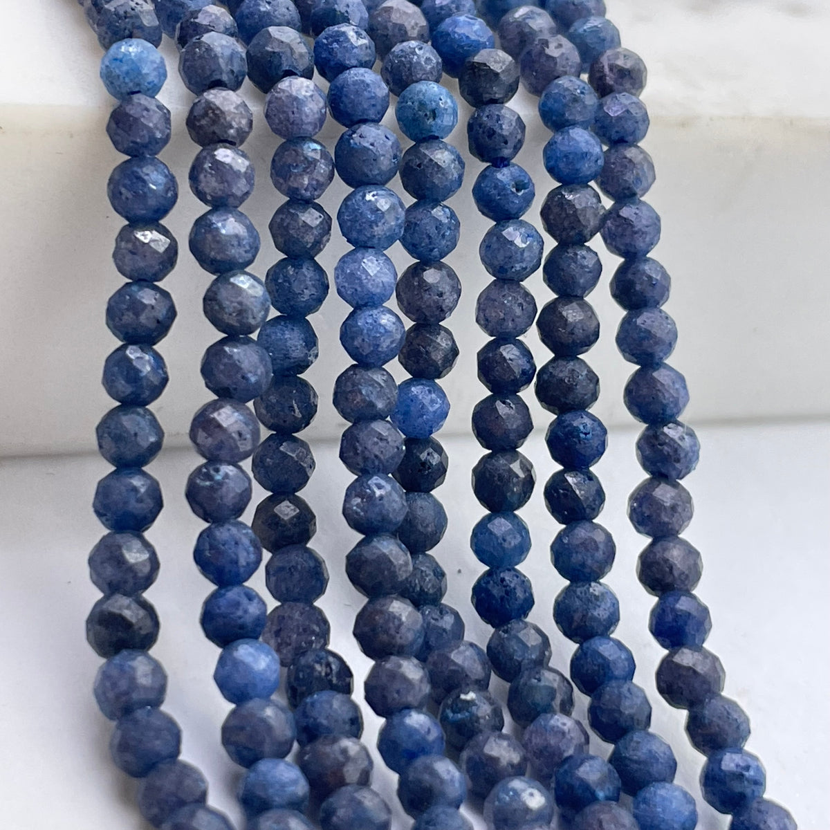 Faceted blue sapphire beads strung in multiple strands, displayed on a white marble-like surface.