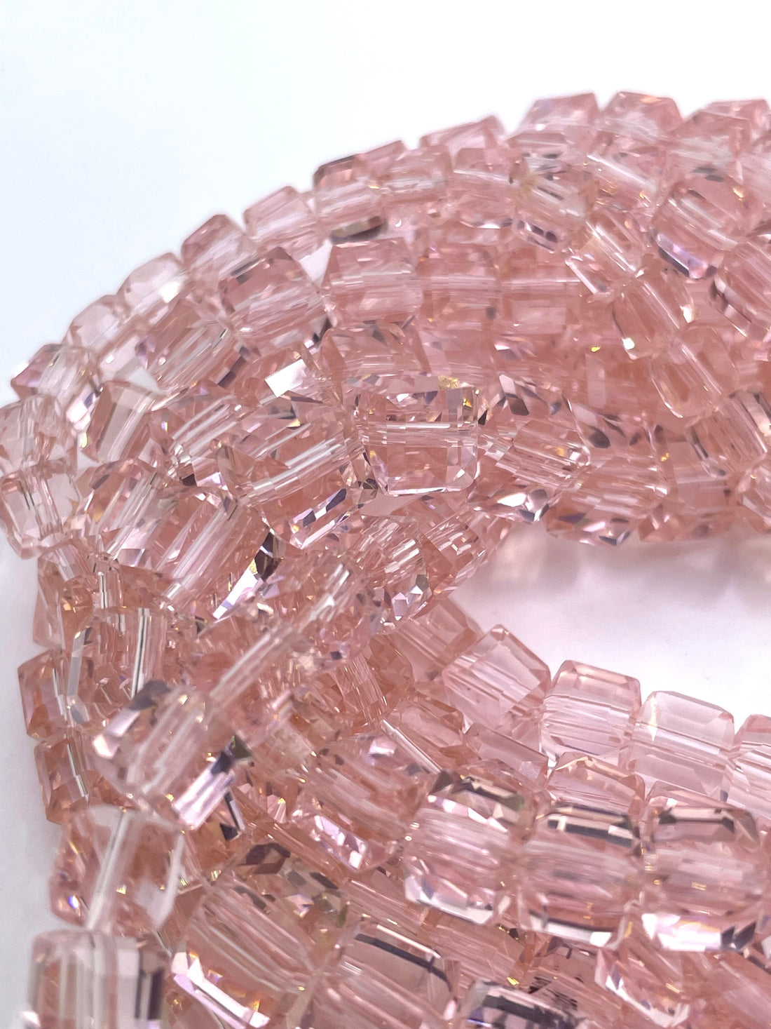 A close-up of a strand of faceted, cube rose pink glass beads arranged in a curved, layered pattern against a white background.