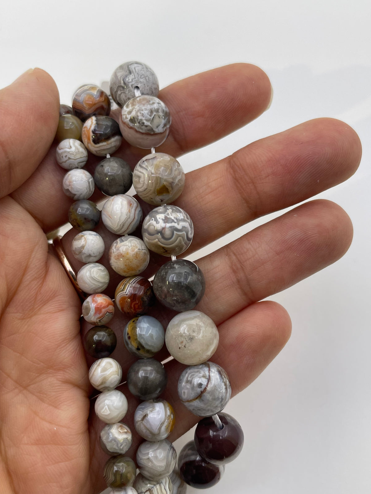 A close-up of a hand holding several strands of round agate stone beads with natural marbled patterns in gray, white, brown, and dark reddish tones against a plain light background.