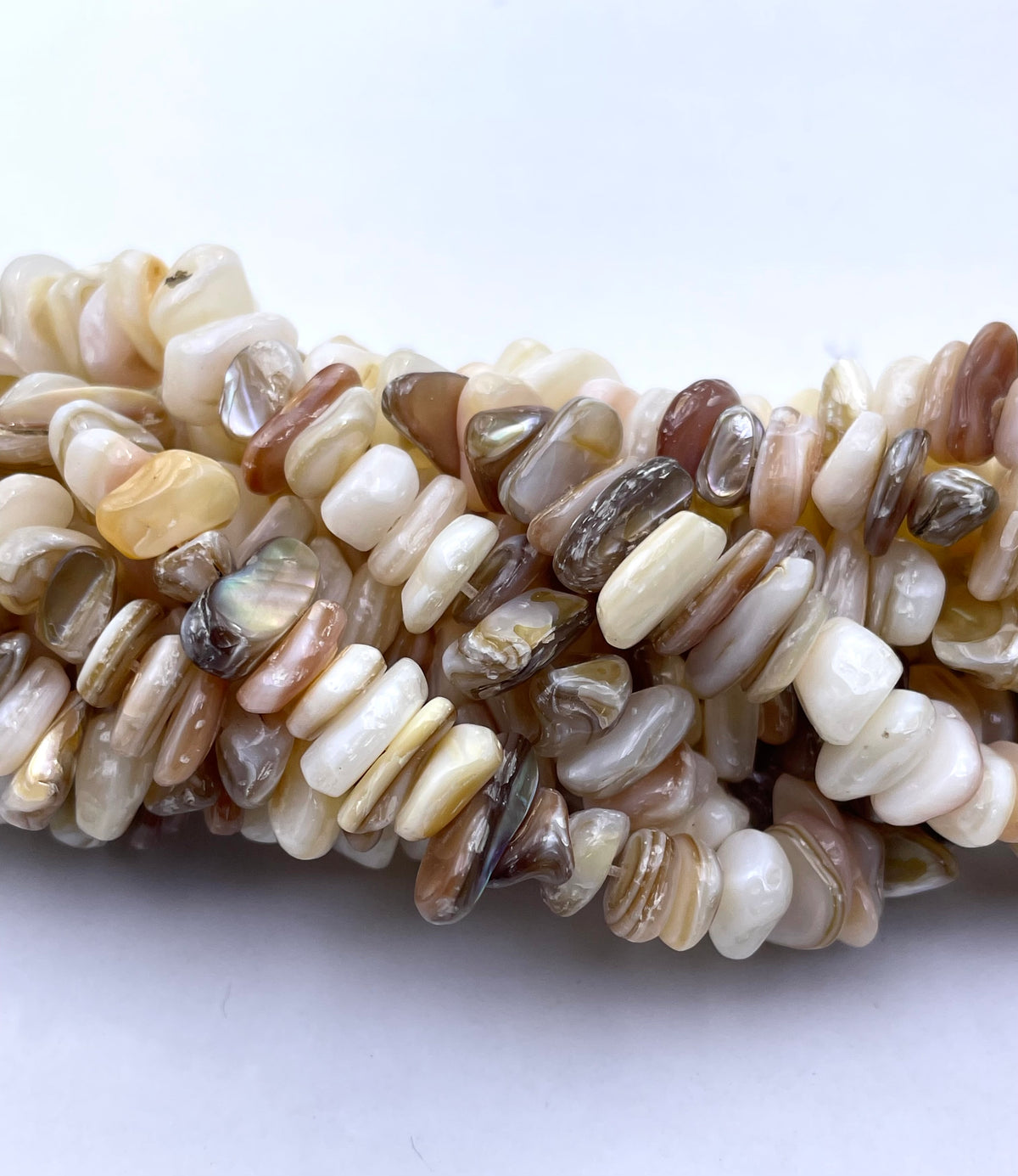 A close-up of a strand of natural stone chips or beads in mixed earthy tones including cream, beige, brown, gray, and translucent milky pieces, arranged horizontally on a plain light background.
