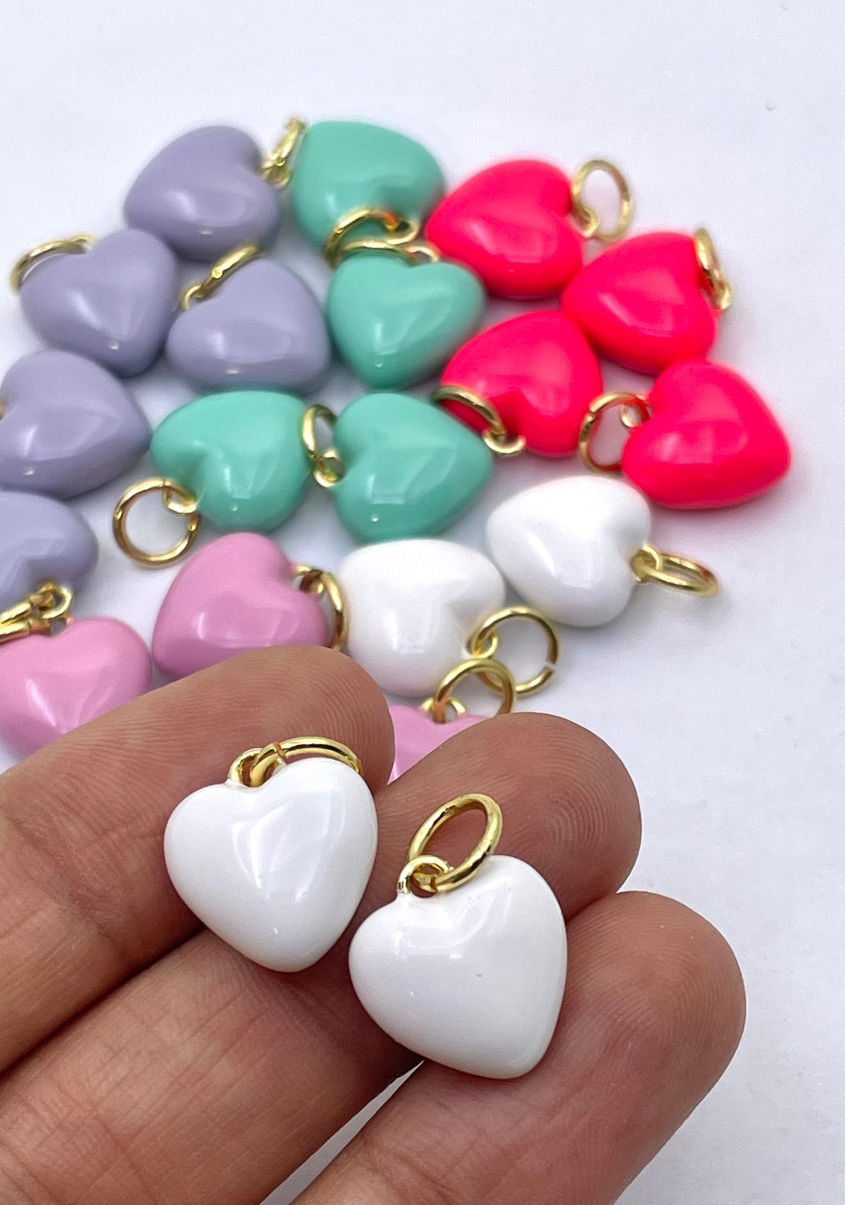 White heart-shaped glossy charms with gold jump rings held in a hand, with other colorful heart charms in pink, purple, mint green, and red scattered in the background on a white surface.