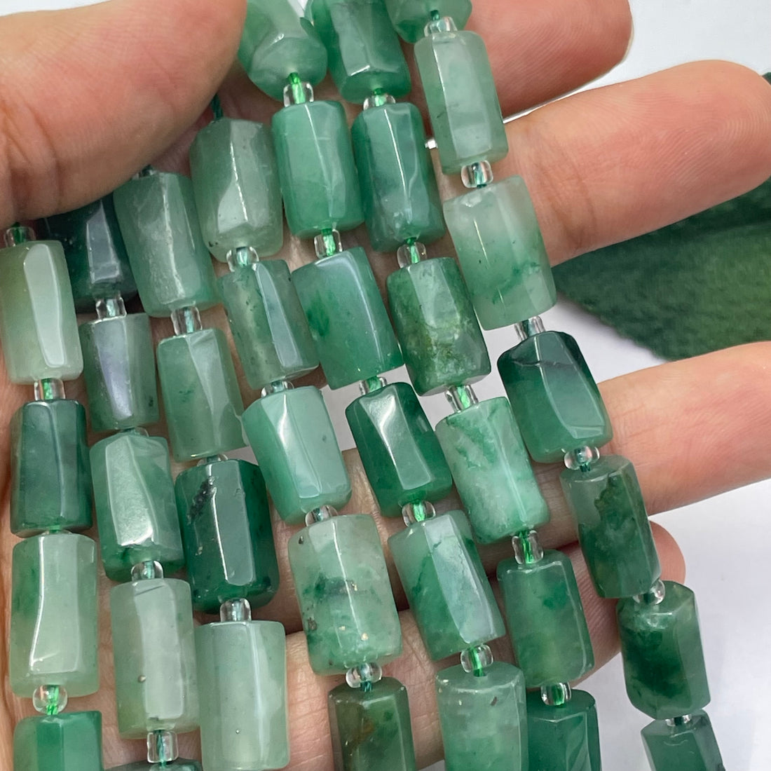 A close-up of a hand holding a strand of rectangular green jade stone beads with a polished, faceted finish.