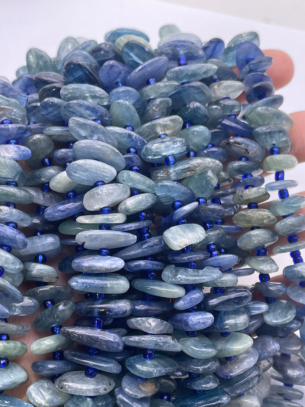 A close-up of a strand of polished blue kyanite beads, strung with small dark blue spacer beads, held in a hand against a white background.