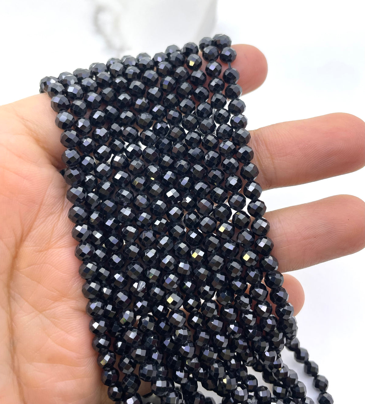 A hand holding several strands of small faceted black spinel beads, against plain light background