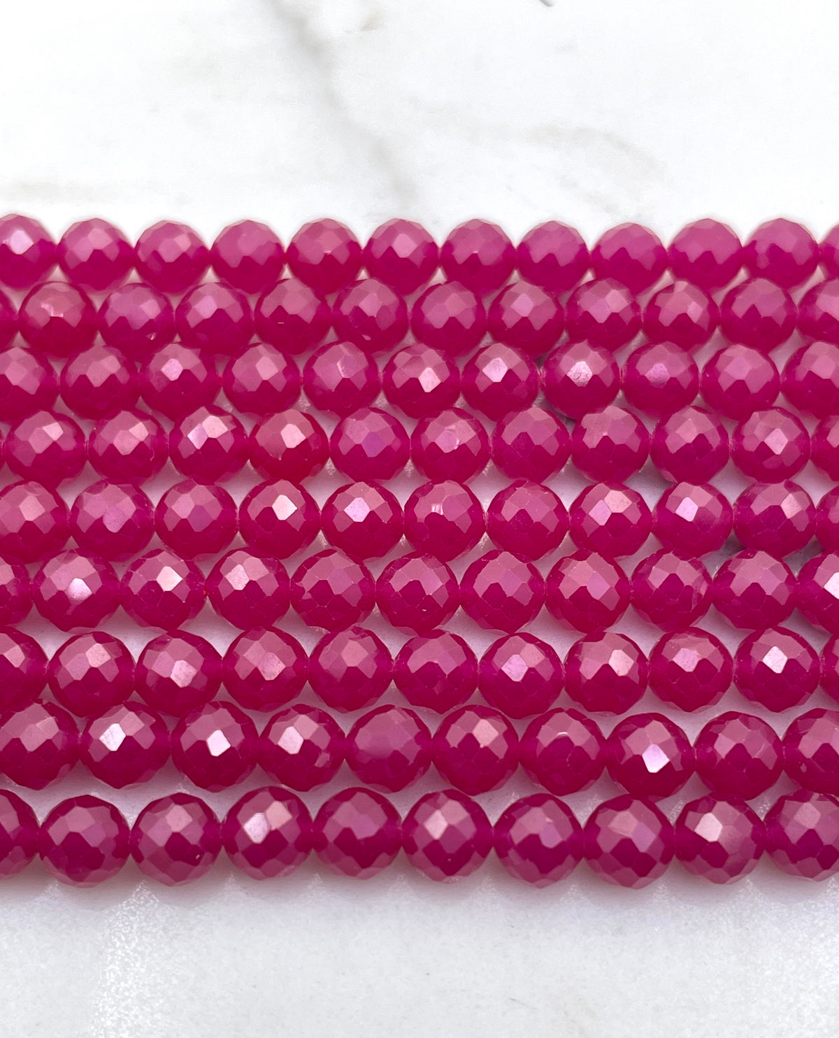 Faceted round ruby beads in a vibrant magenta pink color, arranged in horizontal rows on a white marble surface.