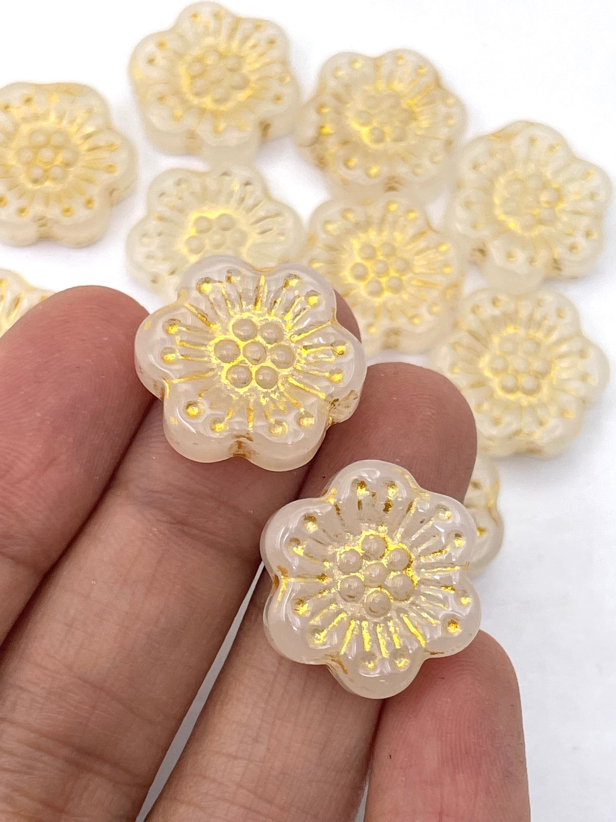 Two small translucent flower-shaped beads with gold foil detailing are held between fingers, with more similar beads blurred in the background on a white surface.