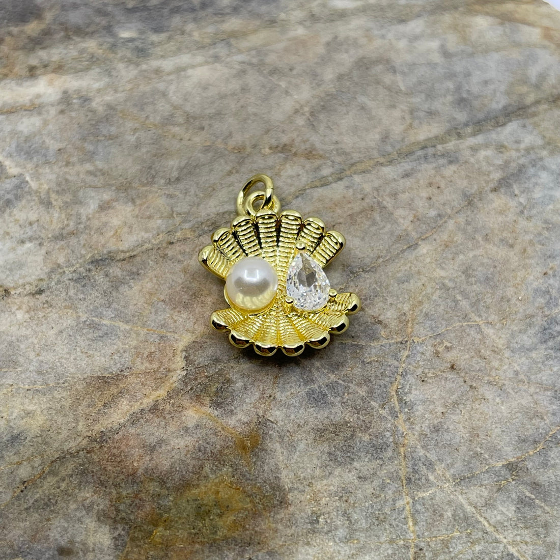 A gold-toned shell-shaped pendant charm featuring a white pearl and a clear teardrop gemstone, displayed on a textured gray stone surface.