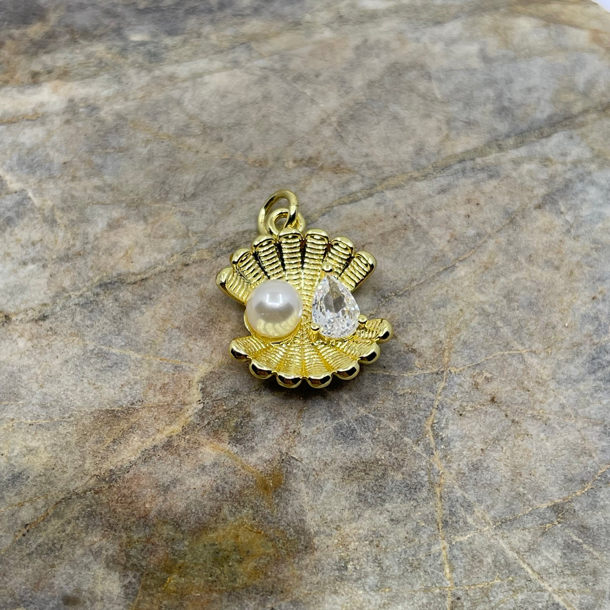 A gold-toned shell-shaped pendant charm featuring a white pearl and a clear teardrop gemstone, displayed on a textured gray stone surface.