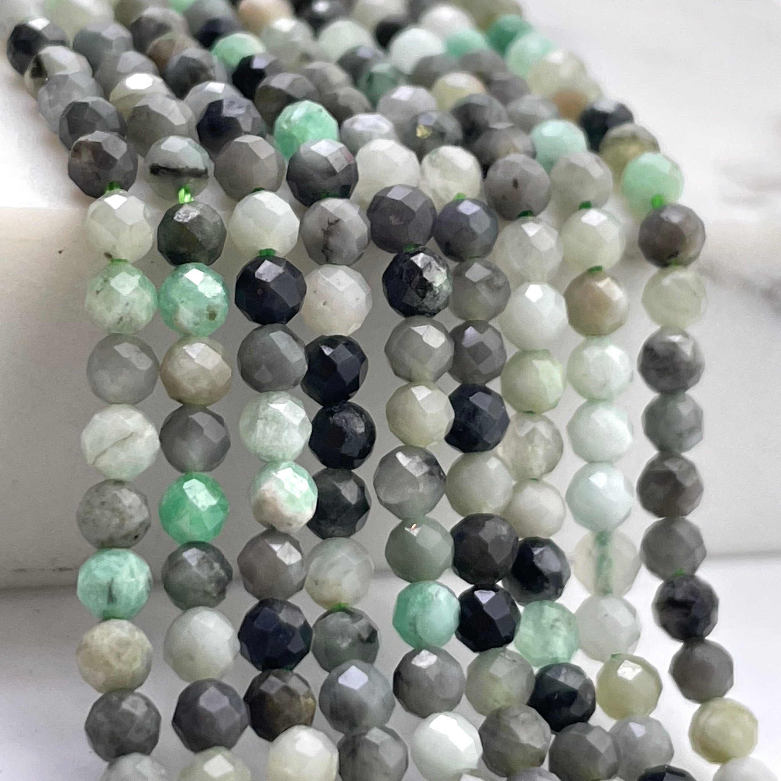 Faceted round emerald gemstone beads in multiple strands, featuring shades of gray, black, white, and light green, arranged on a light marble surface.