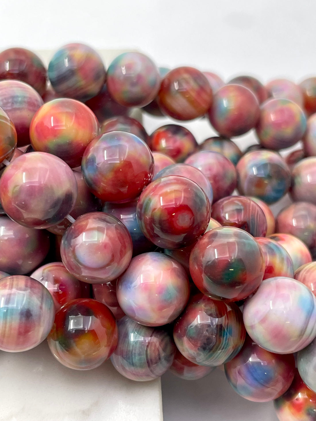 A close-up of glossy round fall foliage agate beads with marbled swirls of pink, red, blue, purple, and white on a white background.