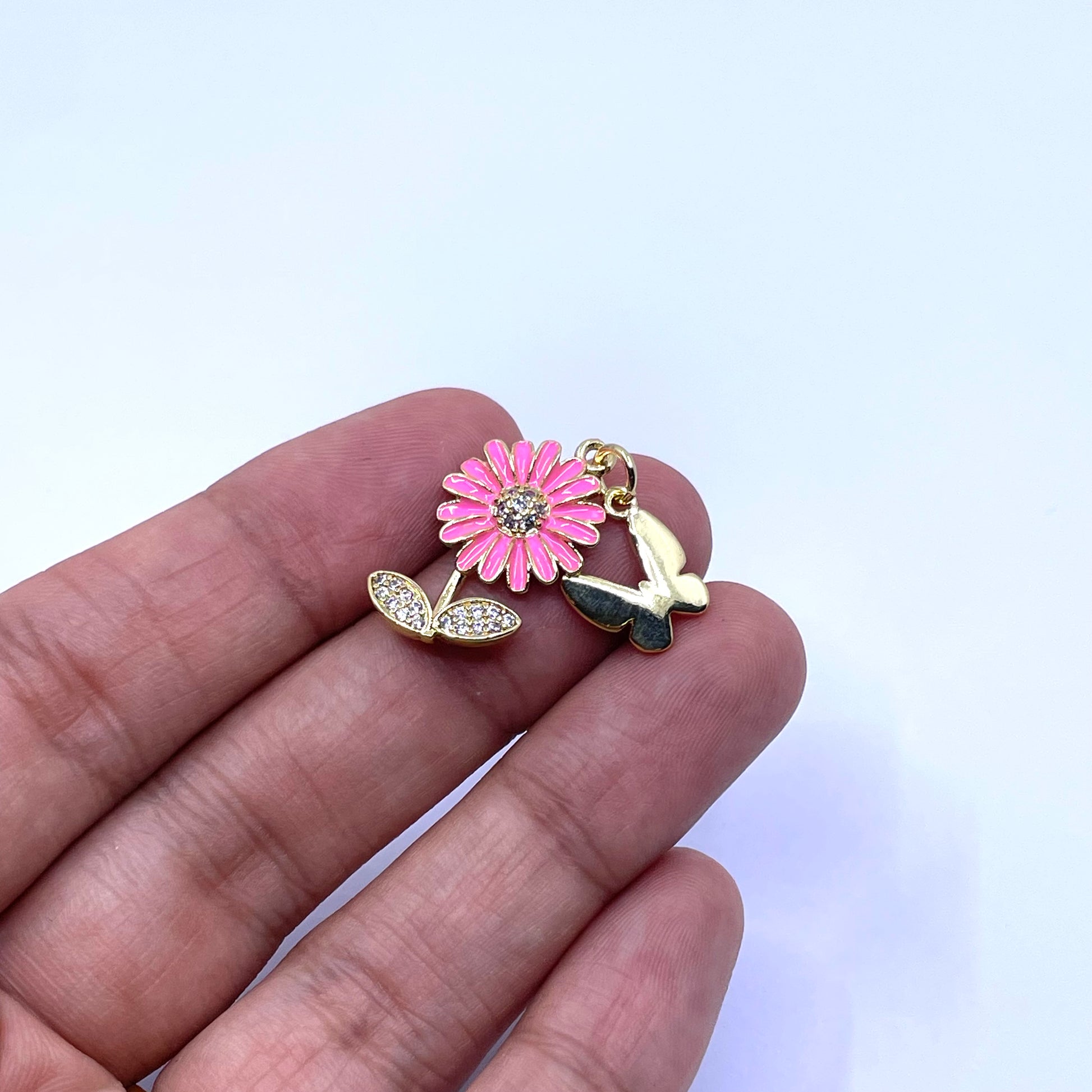 A small jewelry charm or pendant featuring a pink flower with gold-toned metal and a butterfly, shown resting on a person's fingers against a plain light background.