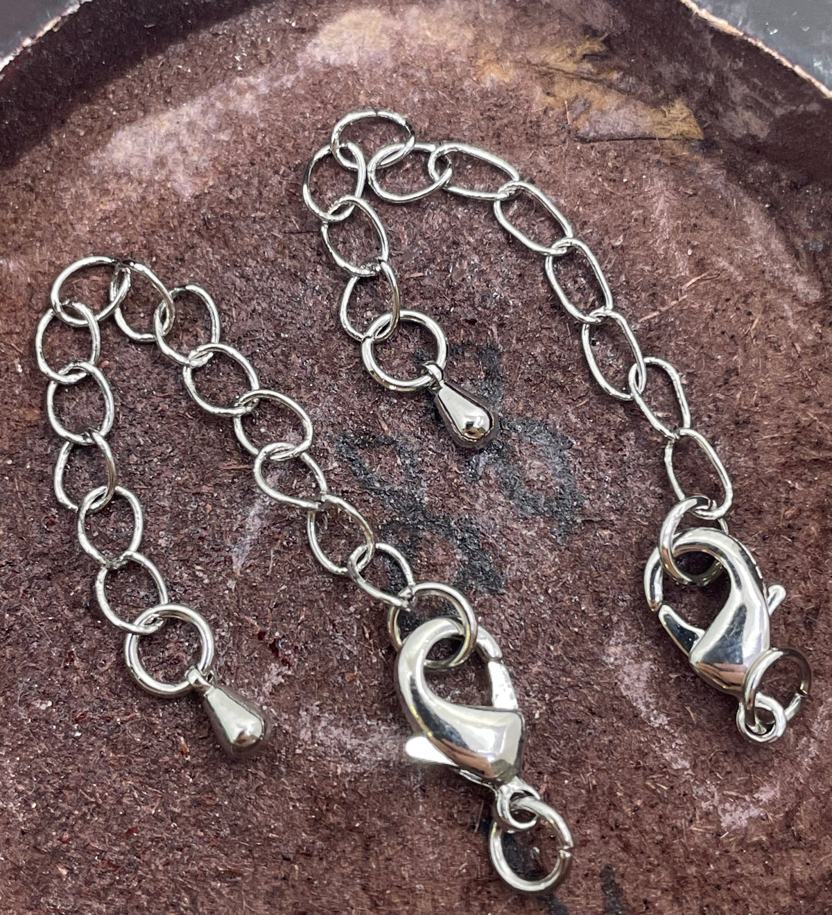 A silver-toned metal chain accessory with lobster clasp ends, photographed on a textured brown surface.