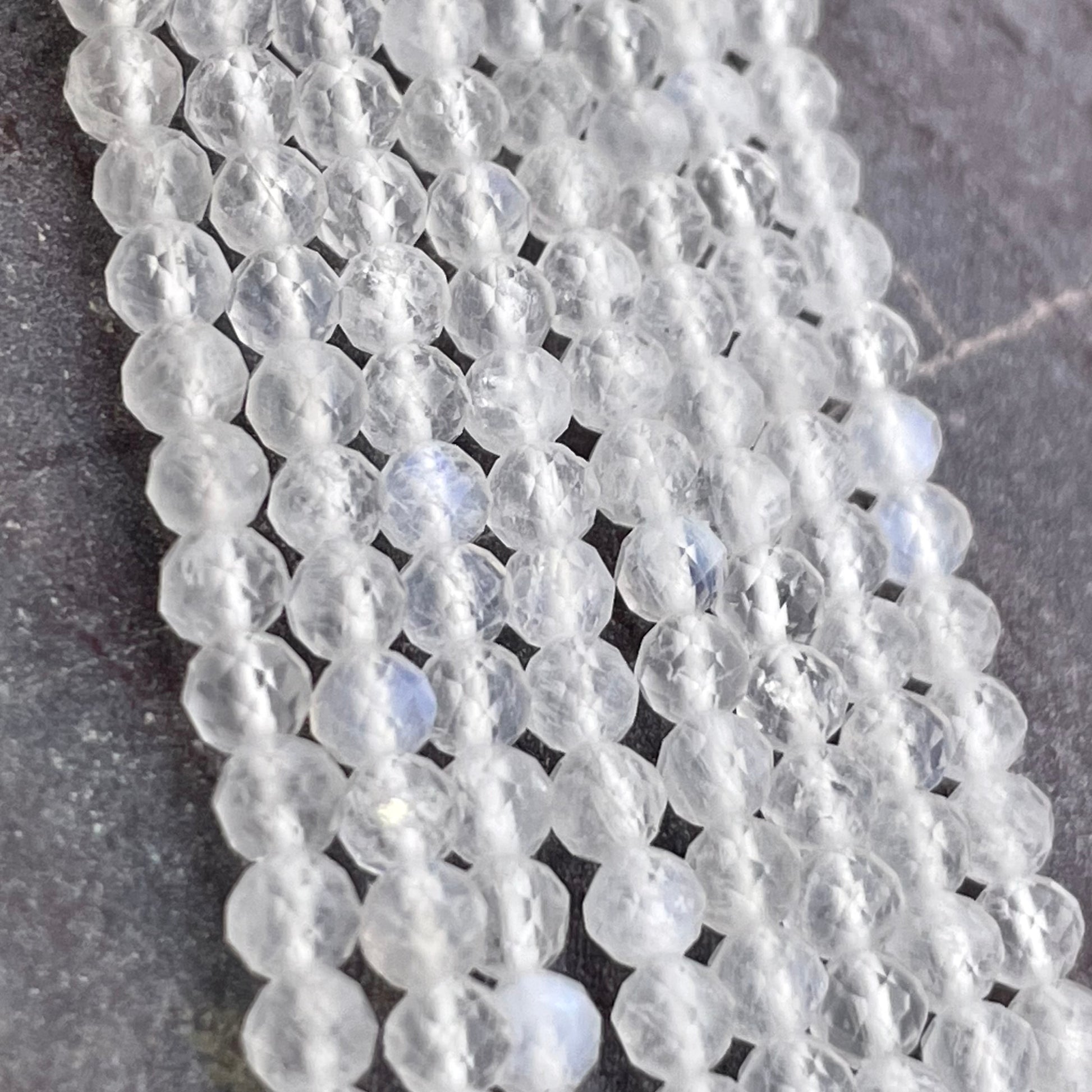 Faceted round moonstone beads in a milky white or clear color, strung in multiple parallel strands on a dark gray textured surface.