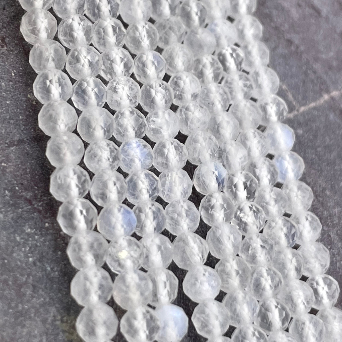 Faceted round moonstone beads in a milky white or clear color, strung in multiple parallel strands on a dark gray textured surface.