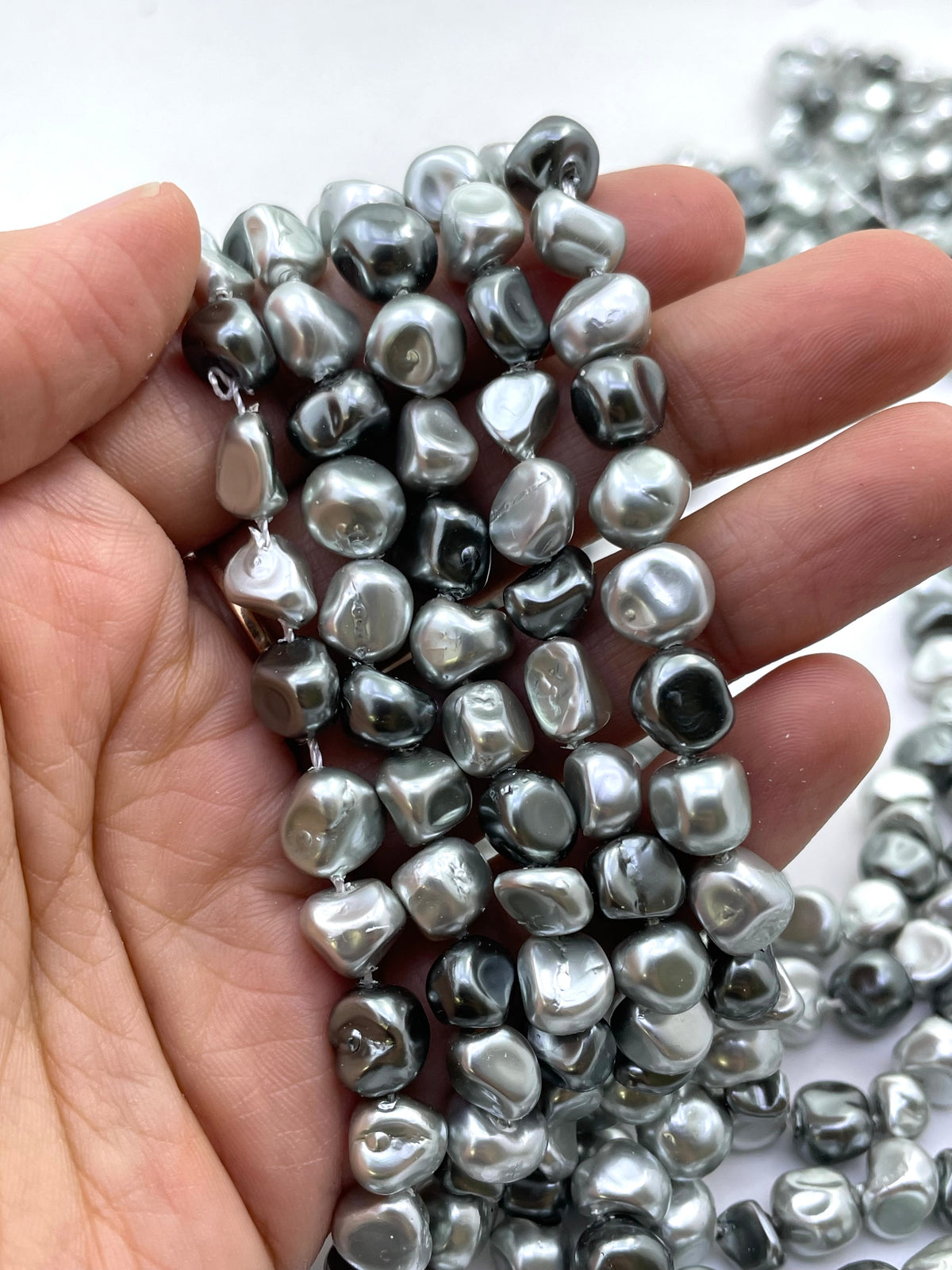 A close-up of a hand holding several strands of irregular, polished silver-gray shell pearls against a white background.
