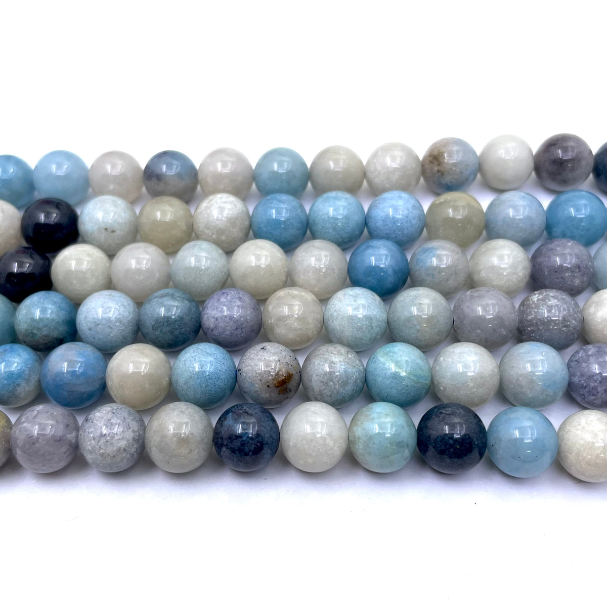 Multiple strands of round polished trolleite gemstone beads in shades of light blue, white, gray, and dark navy, arranged horizontally on a white background.