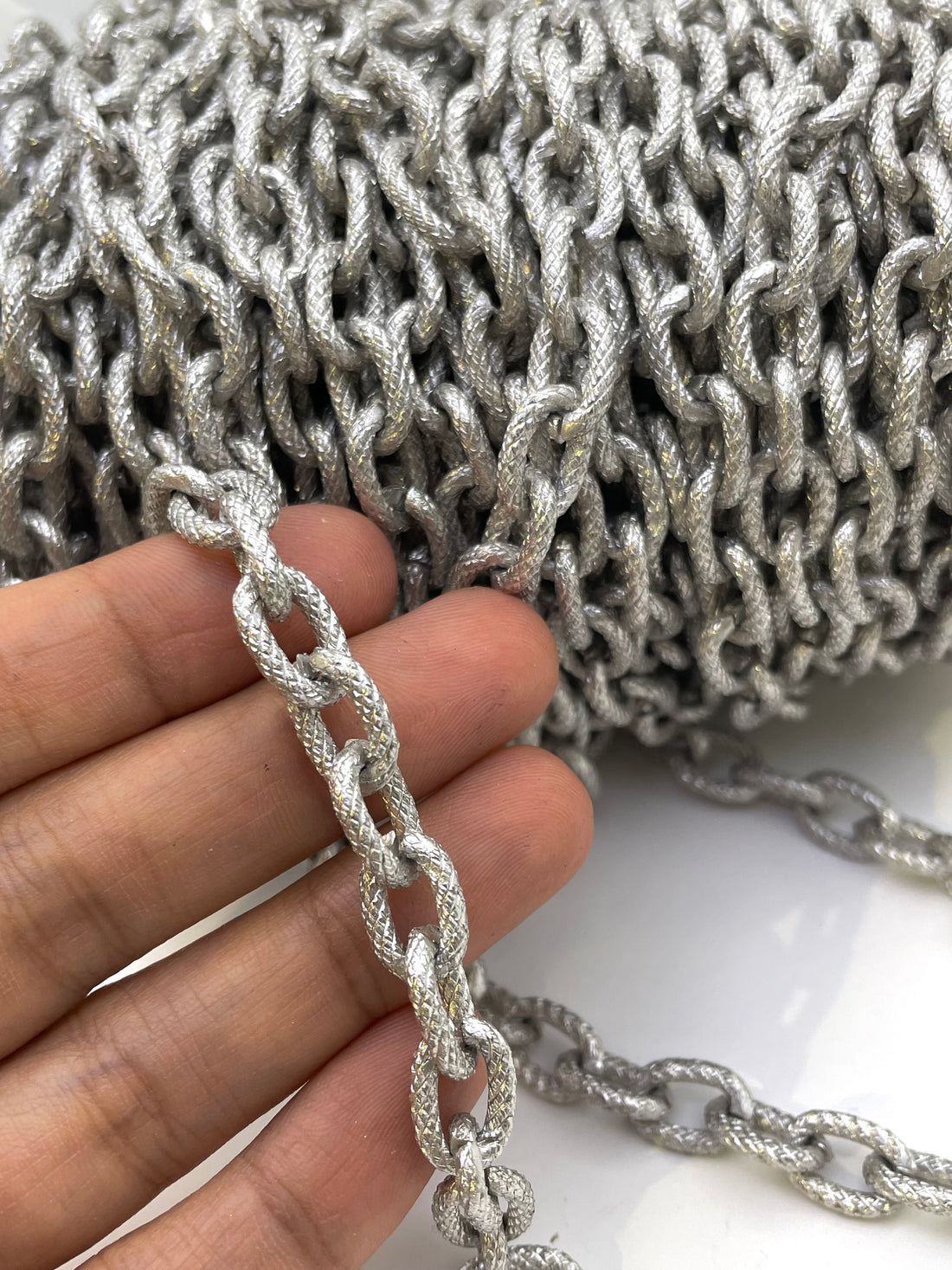 A close-up of a silver metallic chain with textured, rope-like links, shown coiled in the background and draped in the foreground on a white surface.