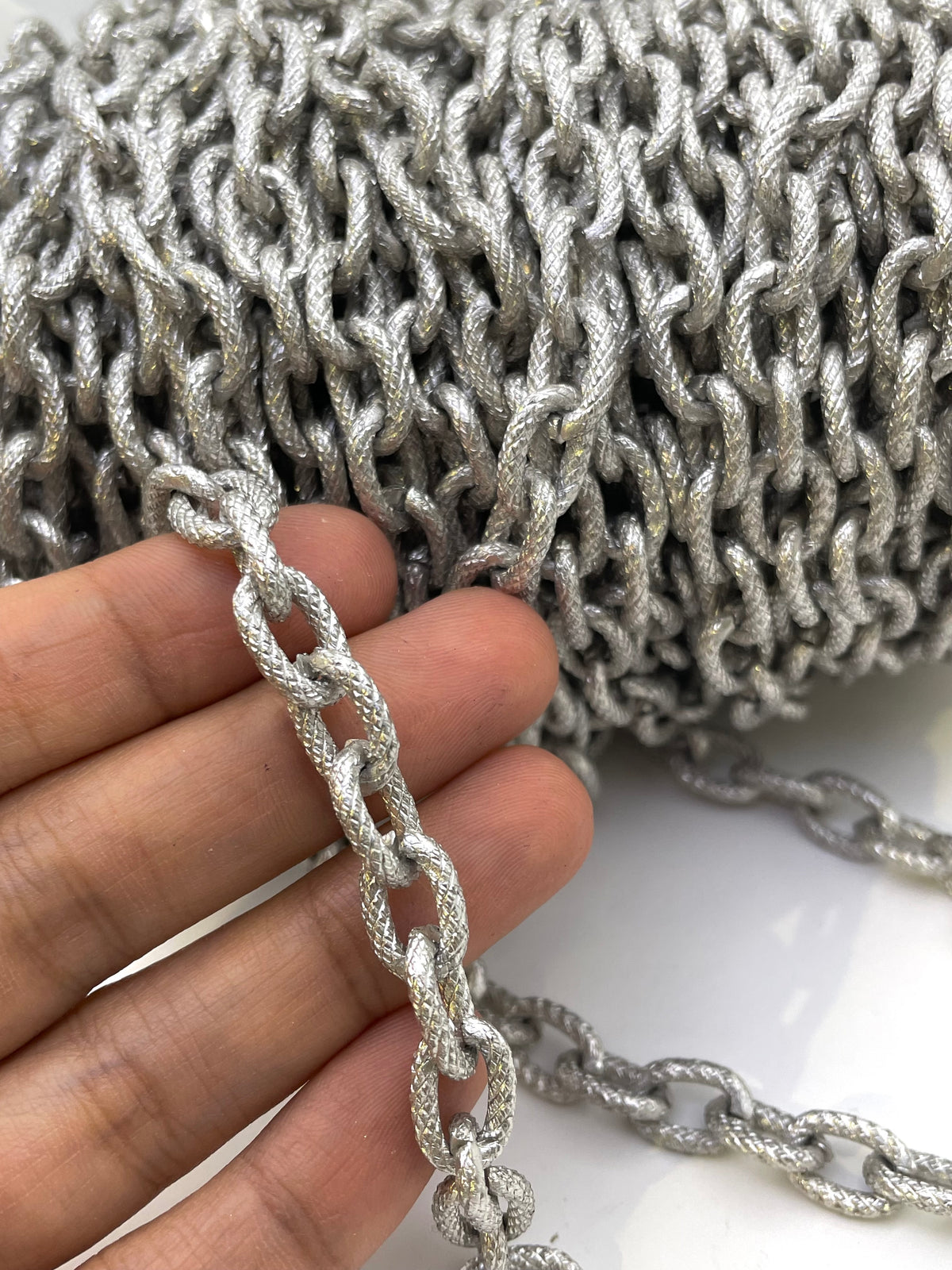 A close-up of a hand holding a metallic silver chain link, with a large spool of the same silver chain visible in the background.