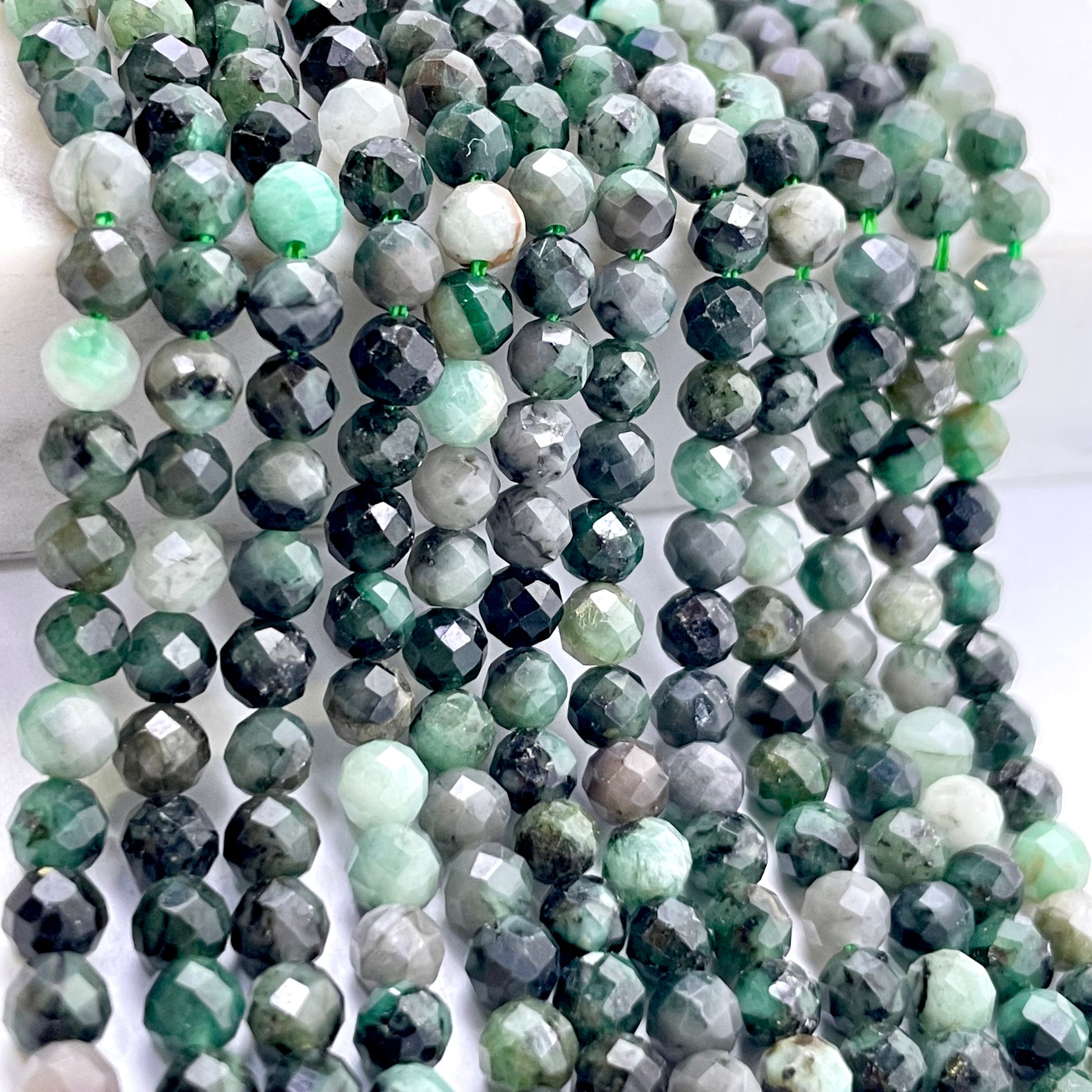 Multiple strands of faceted round emerald beads in shades of green, gray, black, and white, arranged in vertical drapes on a light background.