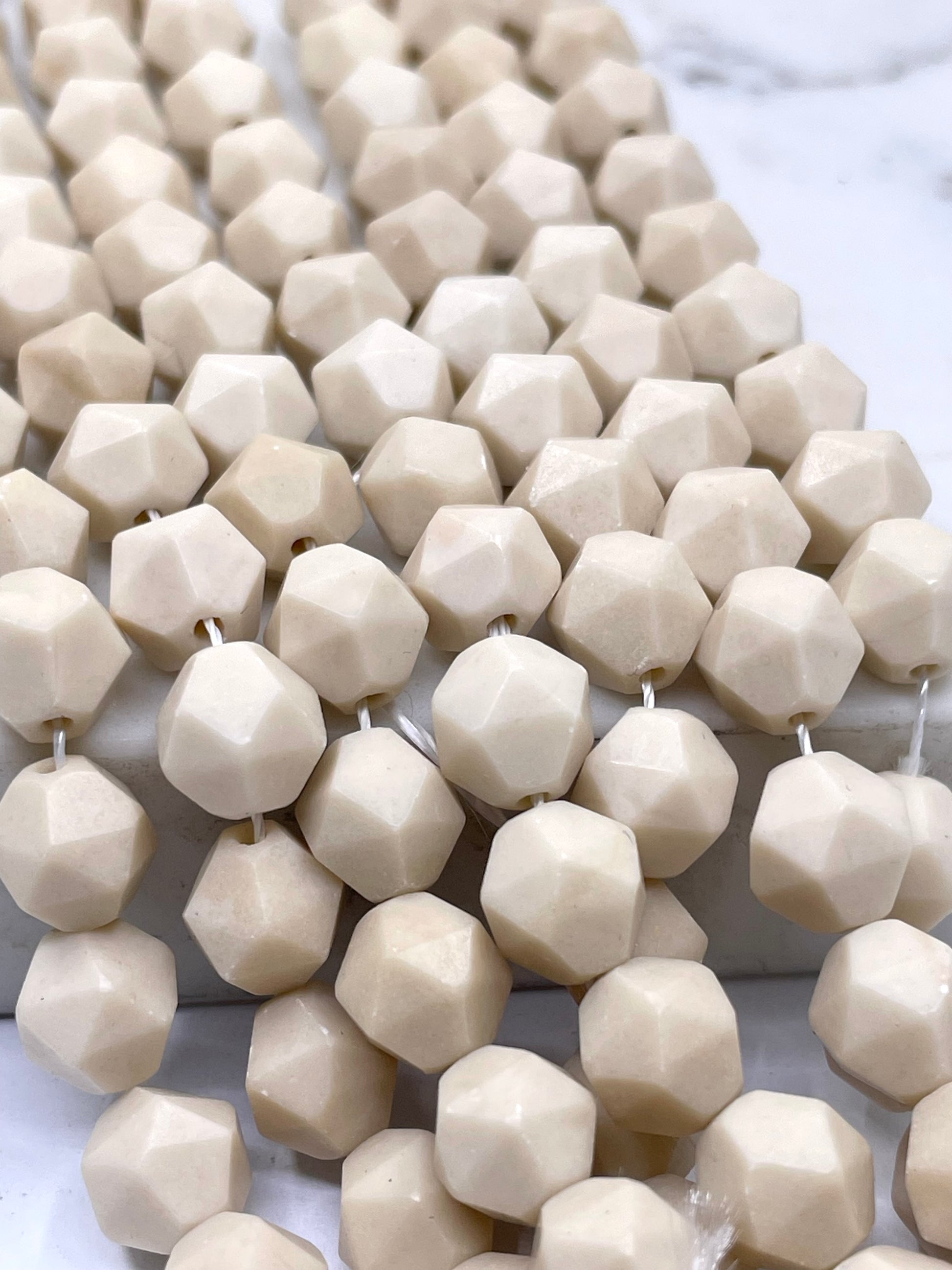 Faceted fossil stone cream-colored beads strung on white cords, arranged diagonally on a light marble surface.