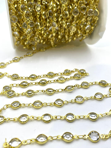 Gold chain trim embellished with clear faceted rhinestones, shown with a spool in the background and several draped strands in the foreground on a white surface.