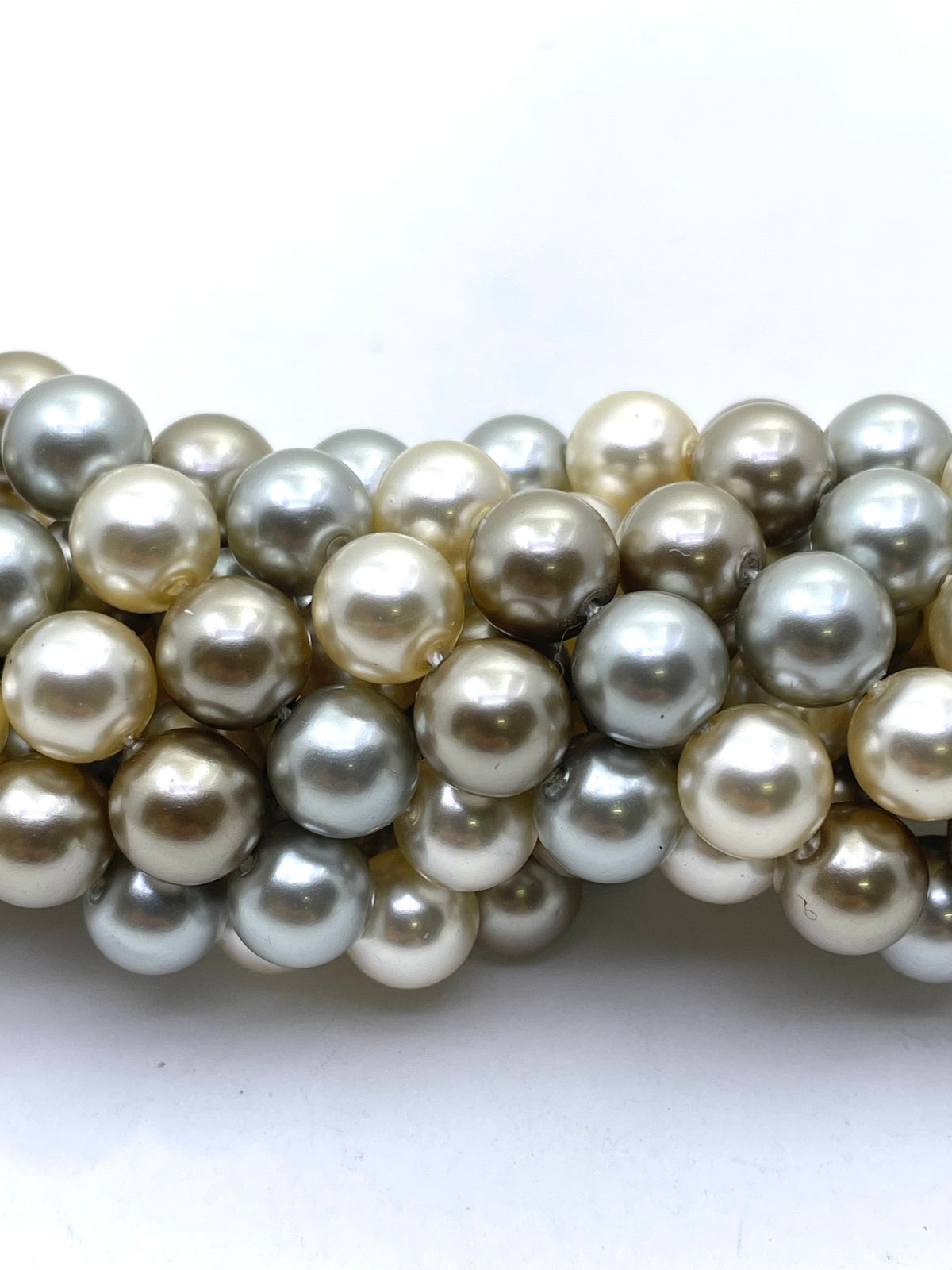 A close-up of a strand of shell pearl beads in mixed natural shades of cream, beige, light brown, and silver-gray, arranged in a curved row on a plain white background.