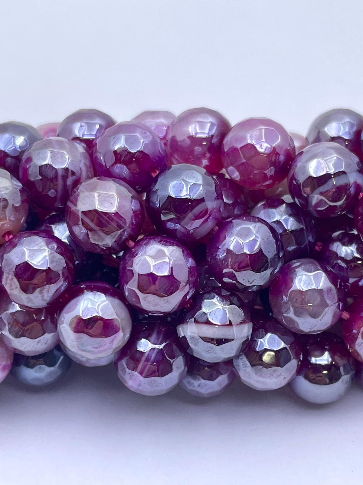 A close-up of faceted round agate beads in a glossy purple and pink gradient, with some beads showing a metallic silver reflective finish, arranged on a plain light background.
