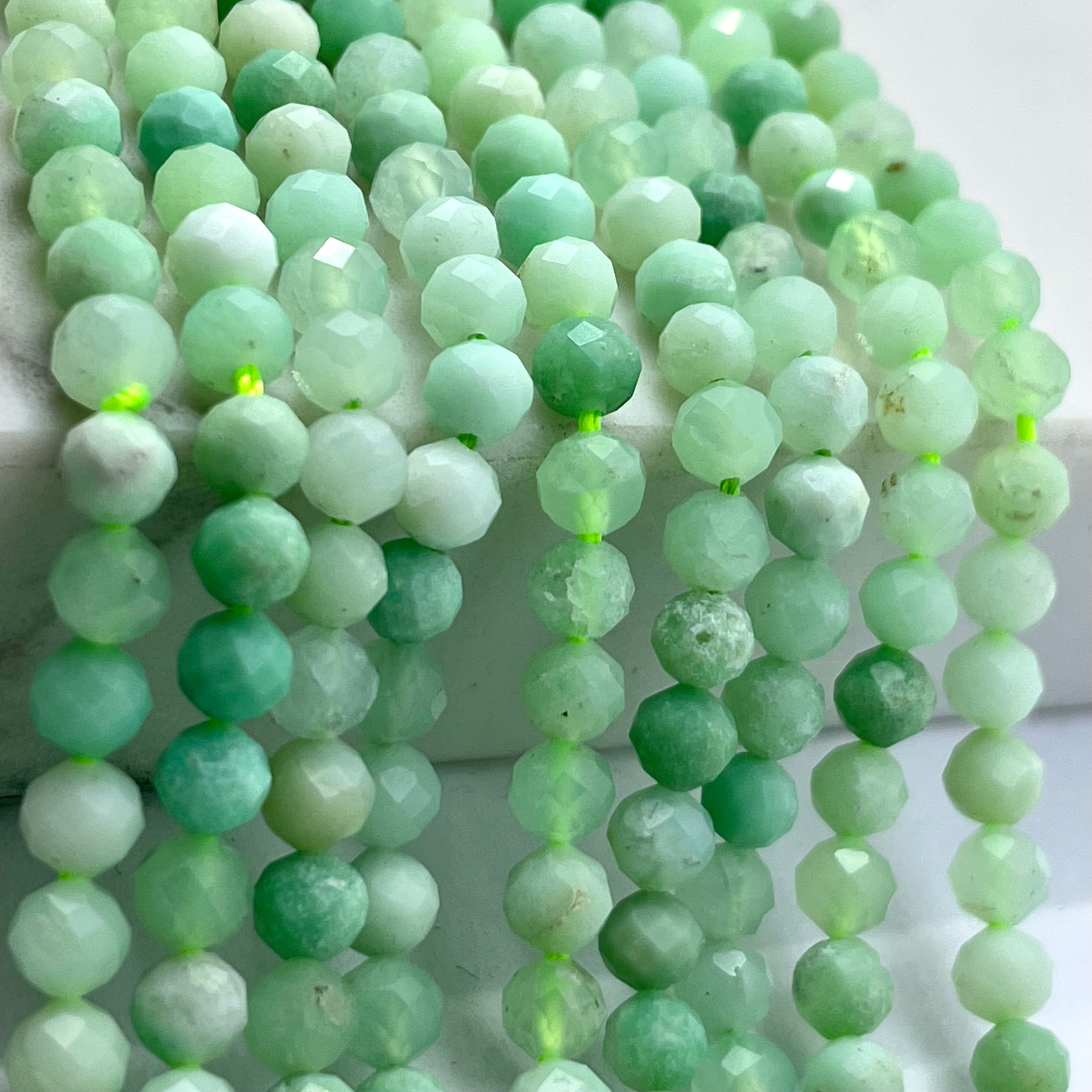 Faceted round chrysoprase gemstone beads in various shades of light green, arranged in strands on a white display block.